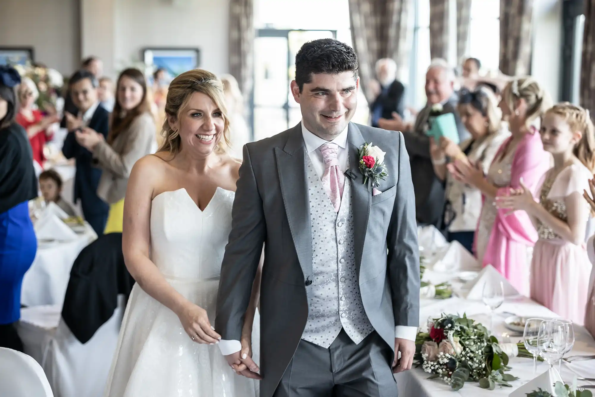 A bride and groom, smiling and holding hands, walk down the aisle of a decorated hall as guests stand and applaud.