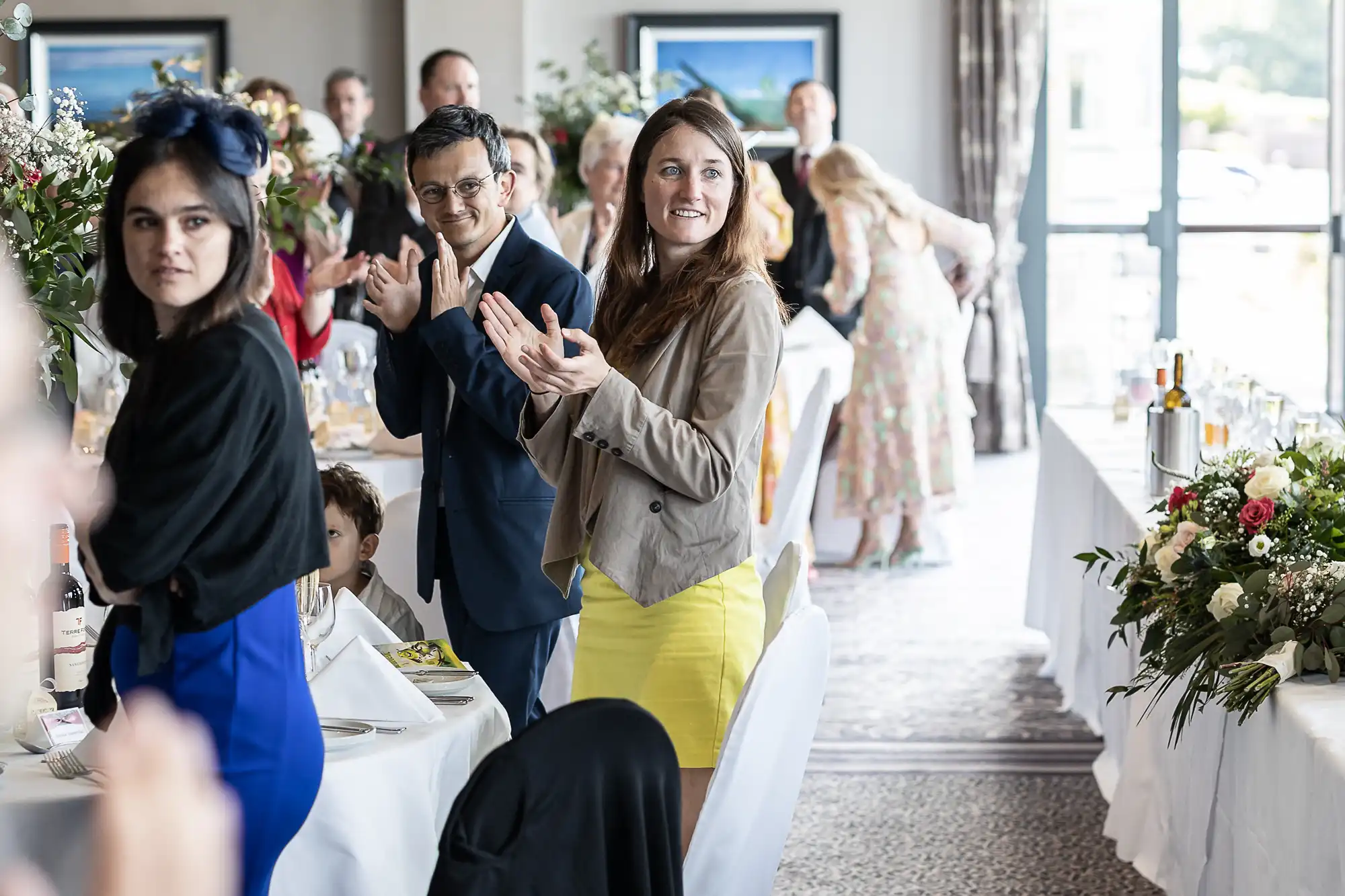 A group of people in a bright room stands and claps, with tables set for a meal and floral arrangements, suggesting a celebration or event.