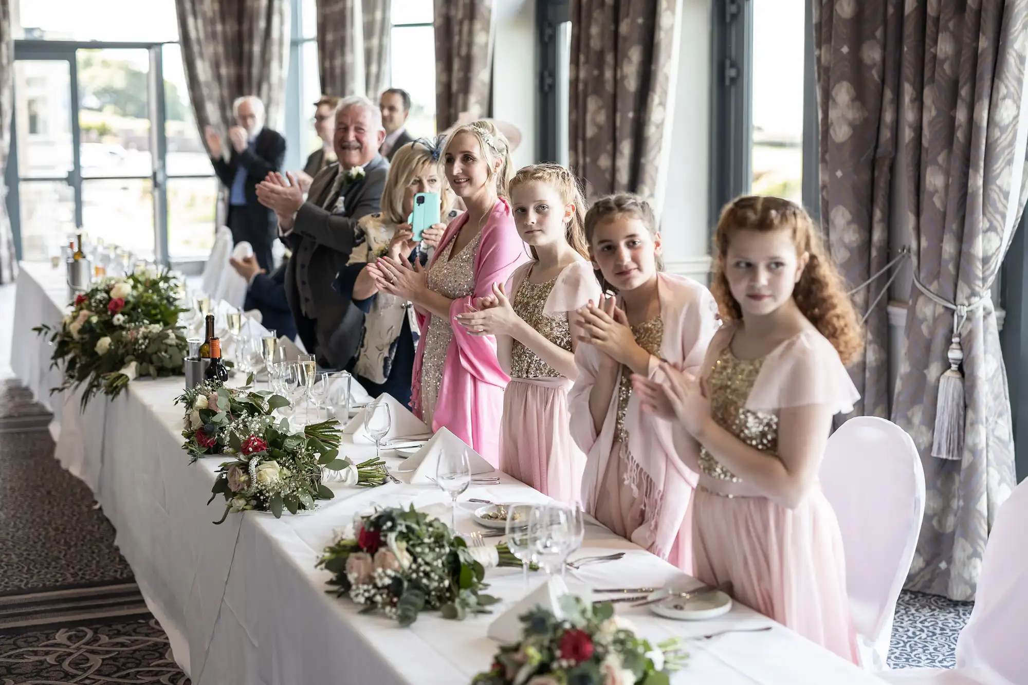 A group of people, including women and girls in matching pink dresses, stand and clap by a long dining table decorated with flowers in a well-lit room with large windows.