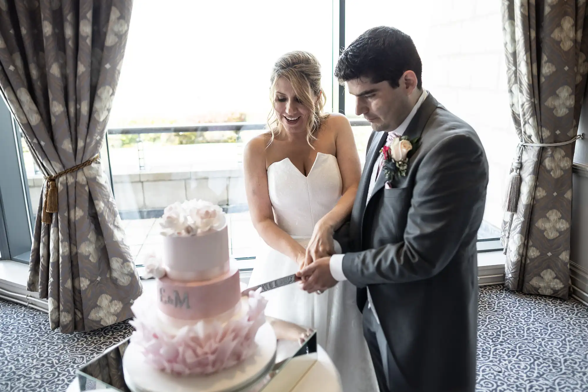 A bride and groom are cutting their wedding cake together. The bride is wearing a white dress, and the groom is in a suit. The cake is pink and white with flowers, and they are in a decorated room.