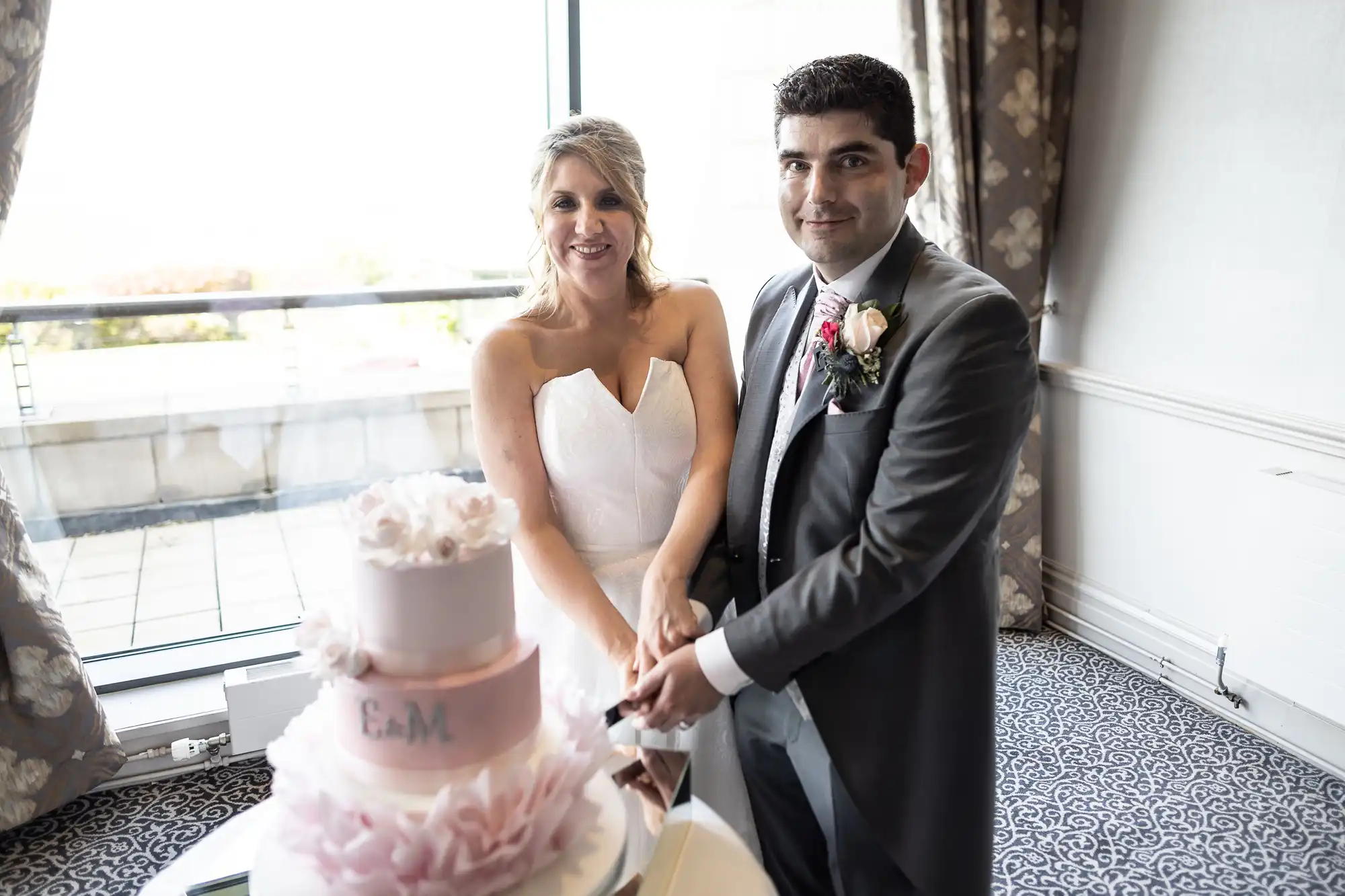 A bride and groom in formal attire are cutting a three-tiered wedding cake with a pink floral design. The couple stands in front of a large window with a floral curtain backdrop.