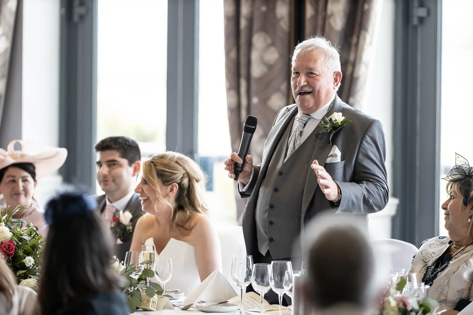 An older man in a suit gives a speech while holding a microphone at a wedding reception. Guests, including the bride and groom, sit at a long table decorated with flowers and place settings.