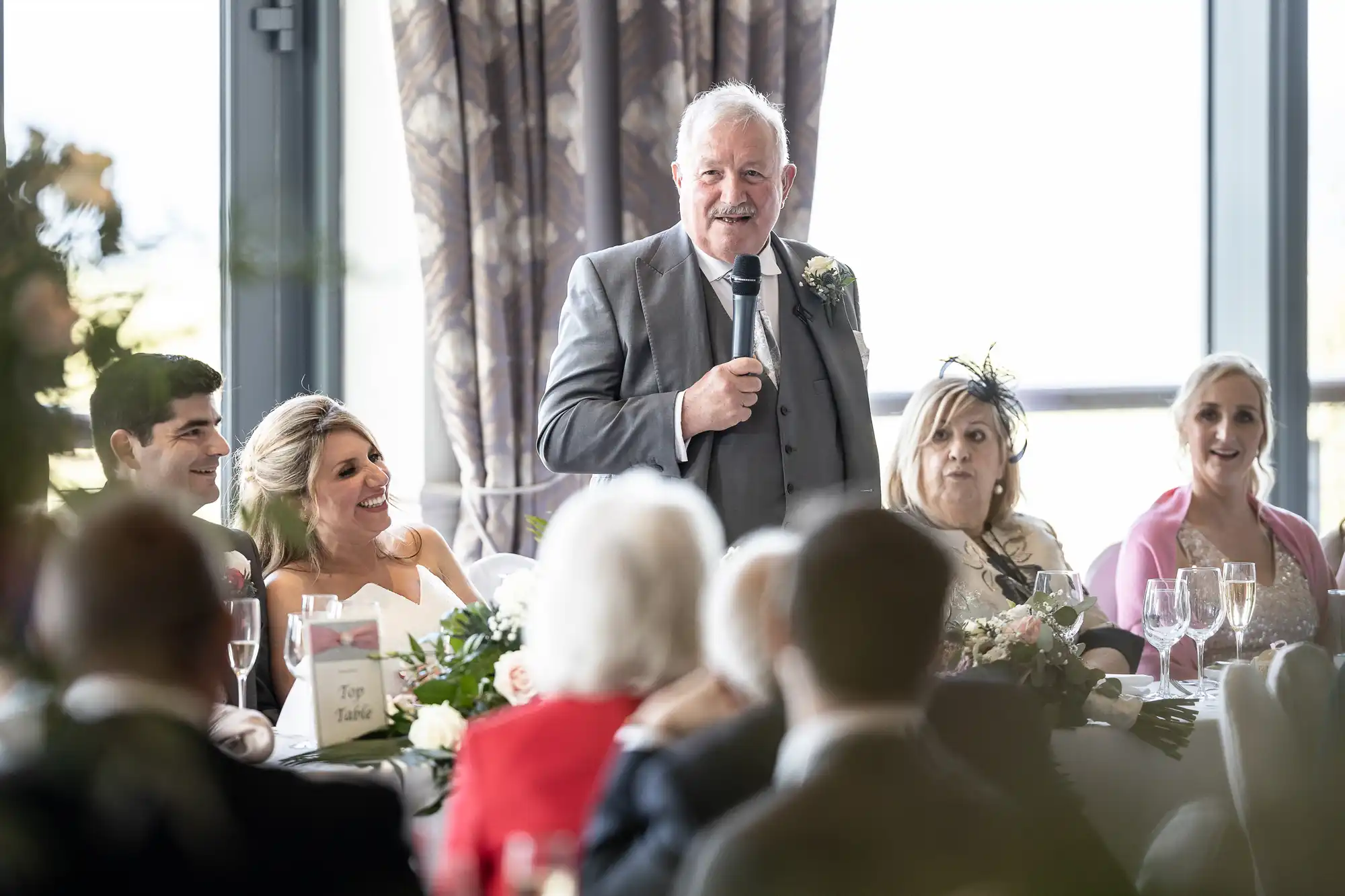 A man in a gray suit is giving a speech with a microphone at a reception, while seated guests listen attentively. A bride and groom are smiling at him from the table.