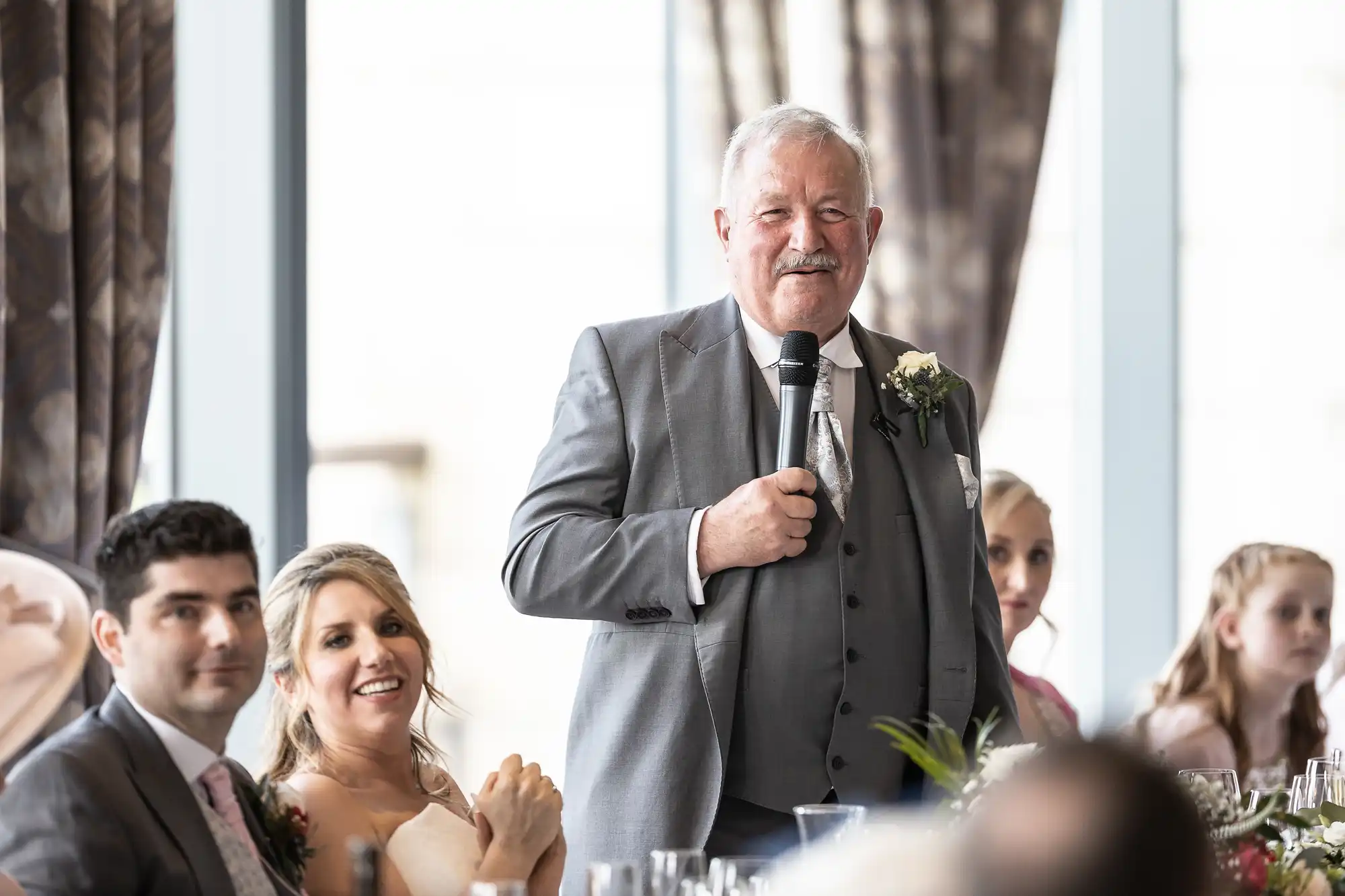 An older man in a grey suit is speaking into a microphone, standing between seated wedding guests at a banquet hall.