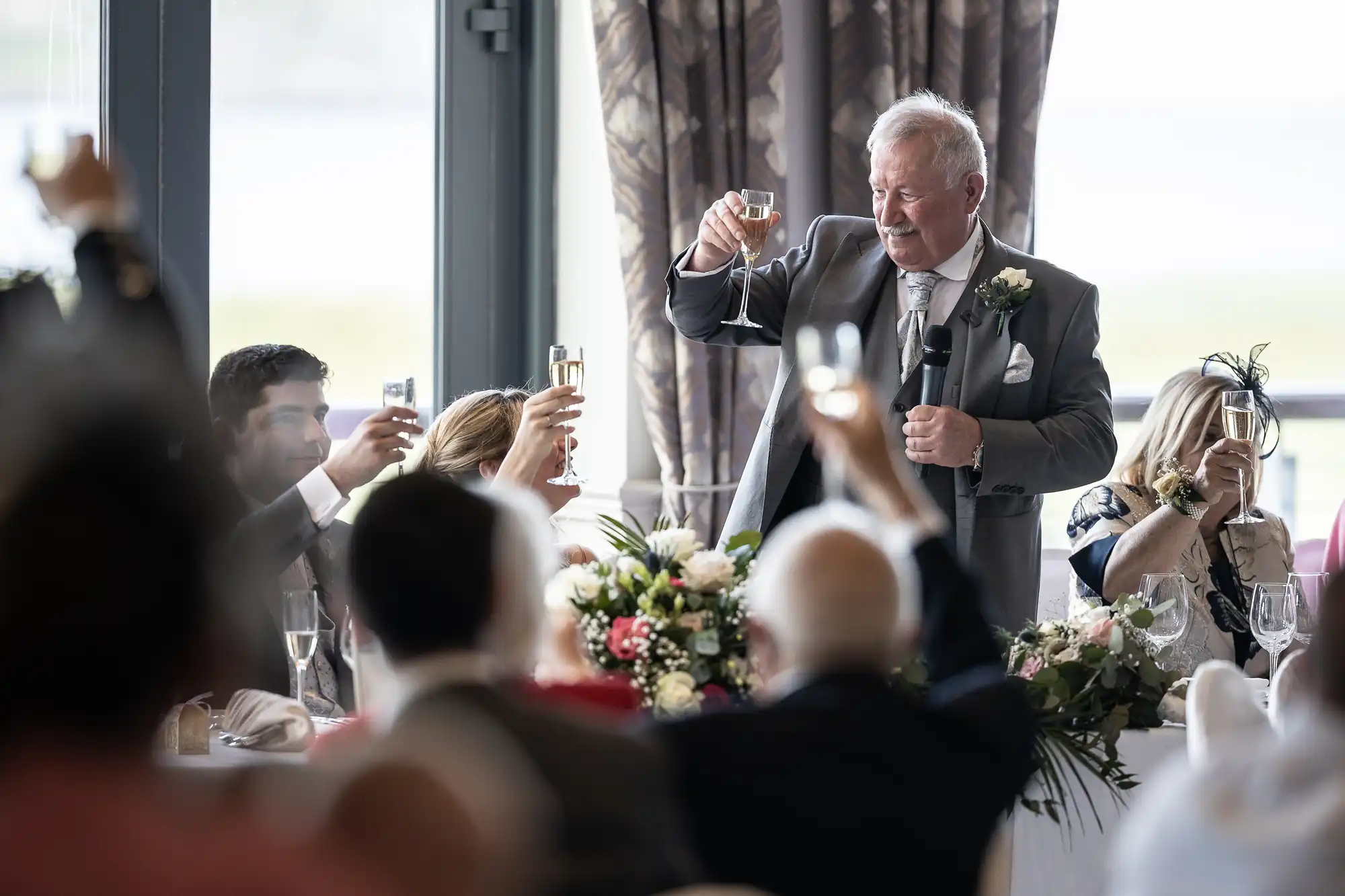 A man in a gray suit holds a microphone and raises a glass for a toast, while seated guests also raise their glasses around a table adorned with floral arrangements.