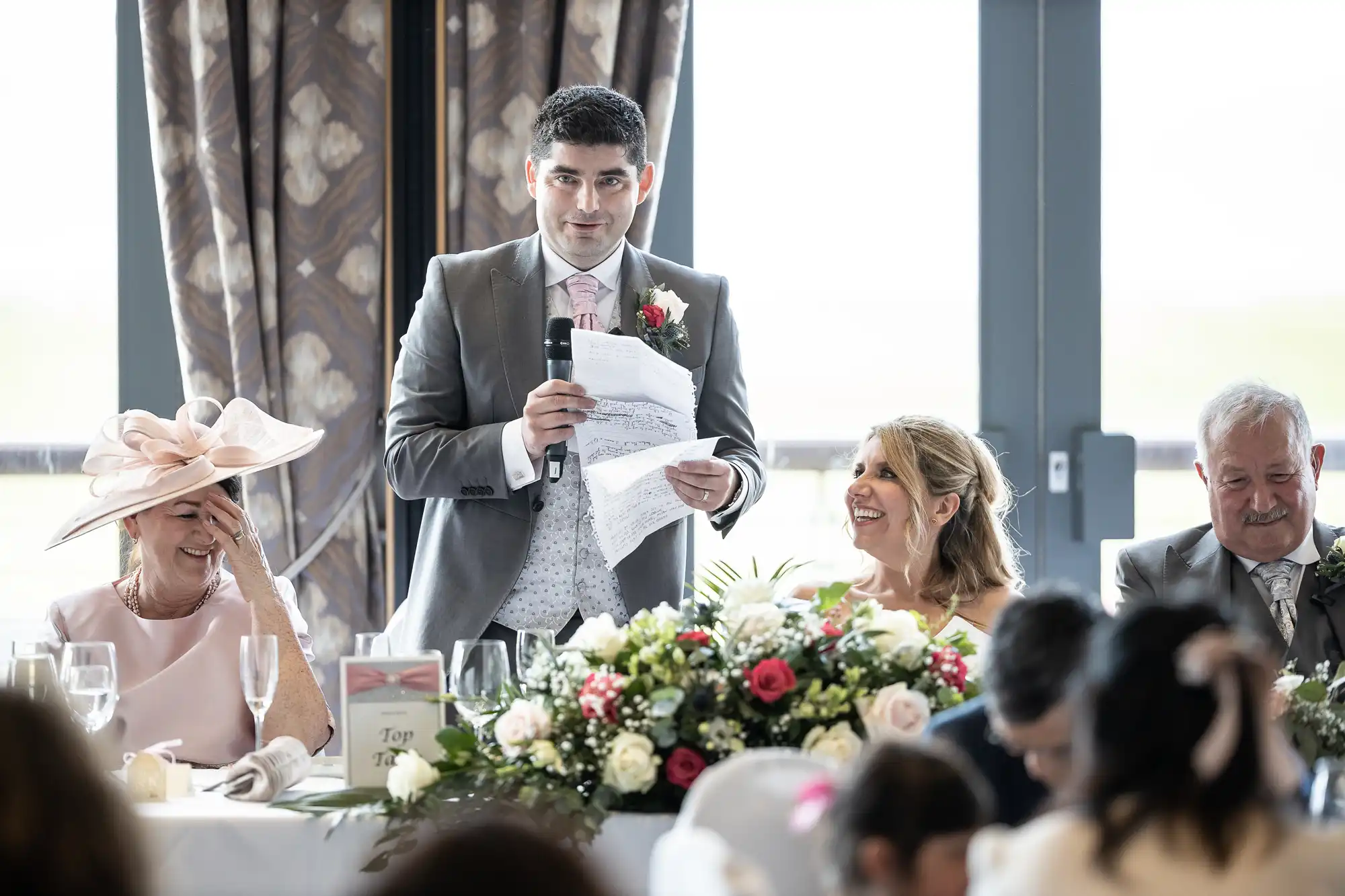 A person in a gray suit and white shirt gives a speech with a microphone in hand, while others seated nearby listen and smile. A table in front is adorned with a bouquet of flowers.