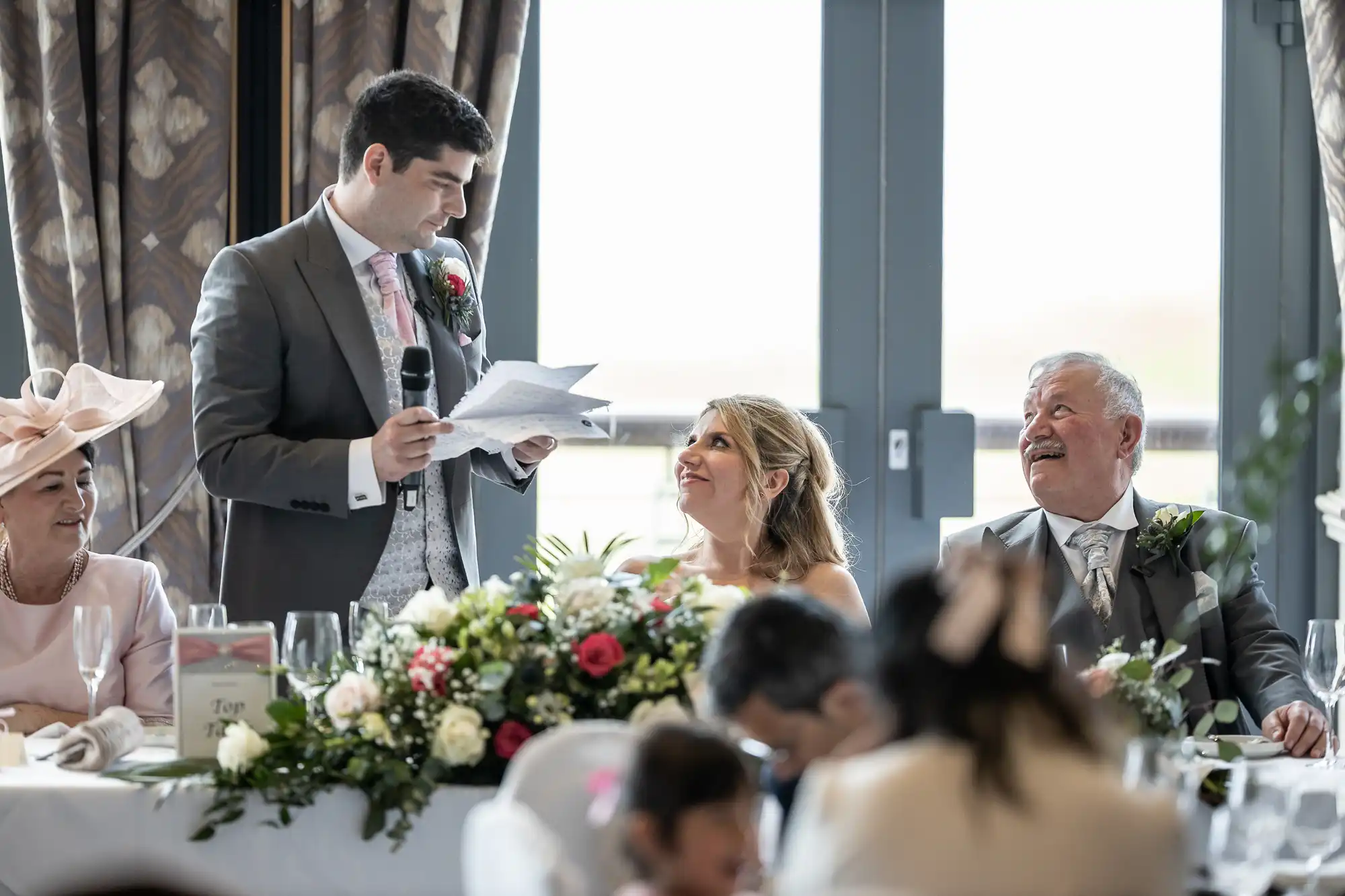 A man in a suit gives a speech with a microphone, standing next to a seated woman in a wedding dress and a man in a suit. A table with flowers and guests are in front of them.