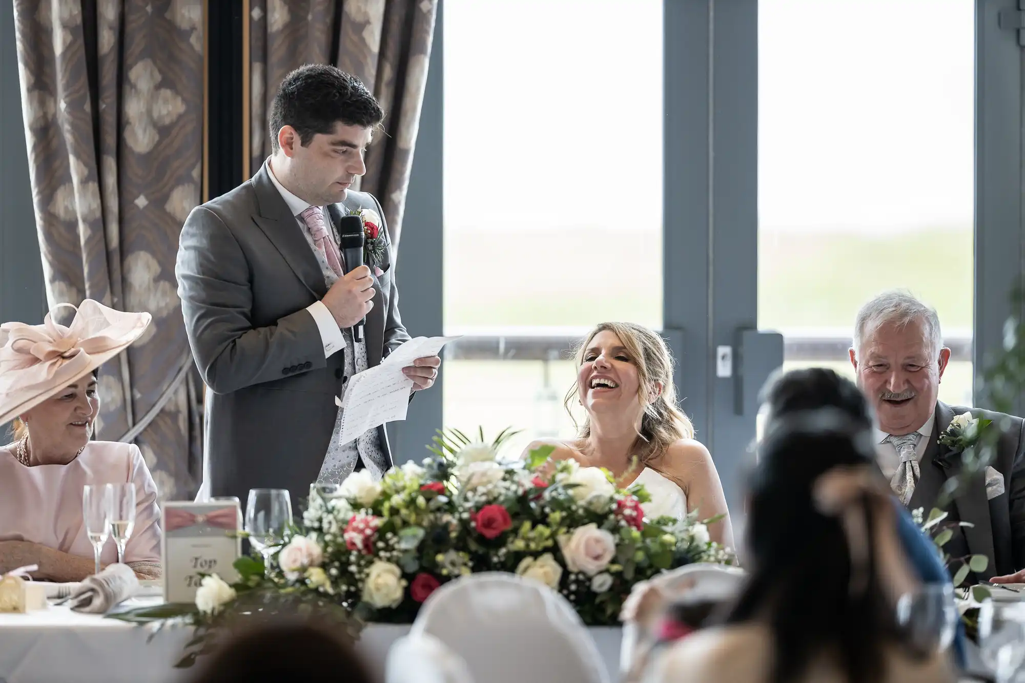 A man in a suit speaks into a microphone at a table, holding a piece of paper. A woman in a white dress laughs, sitting beside him. The table is decorated with a large flower arrangement.