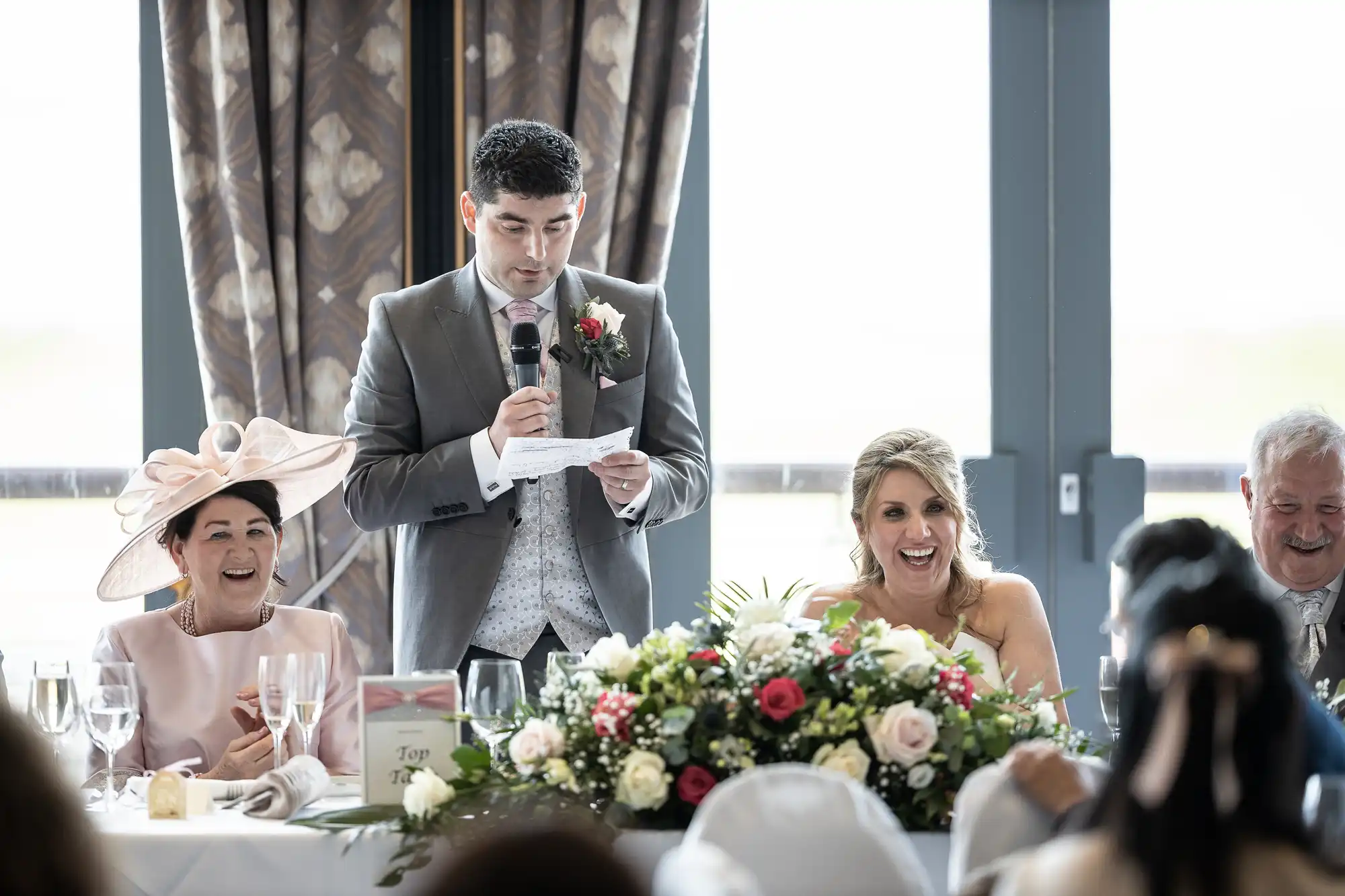 A man in a suit stands and speaks into a microphone, holding a piece of paper. He’s at a table with a floral centerpiece and seated guests, including a woman in a large hat and a smiling woman in a wedding dress.