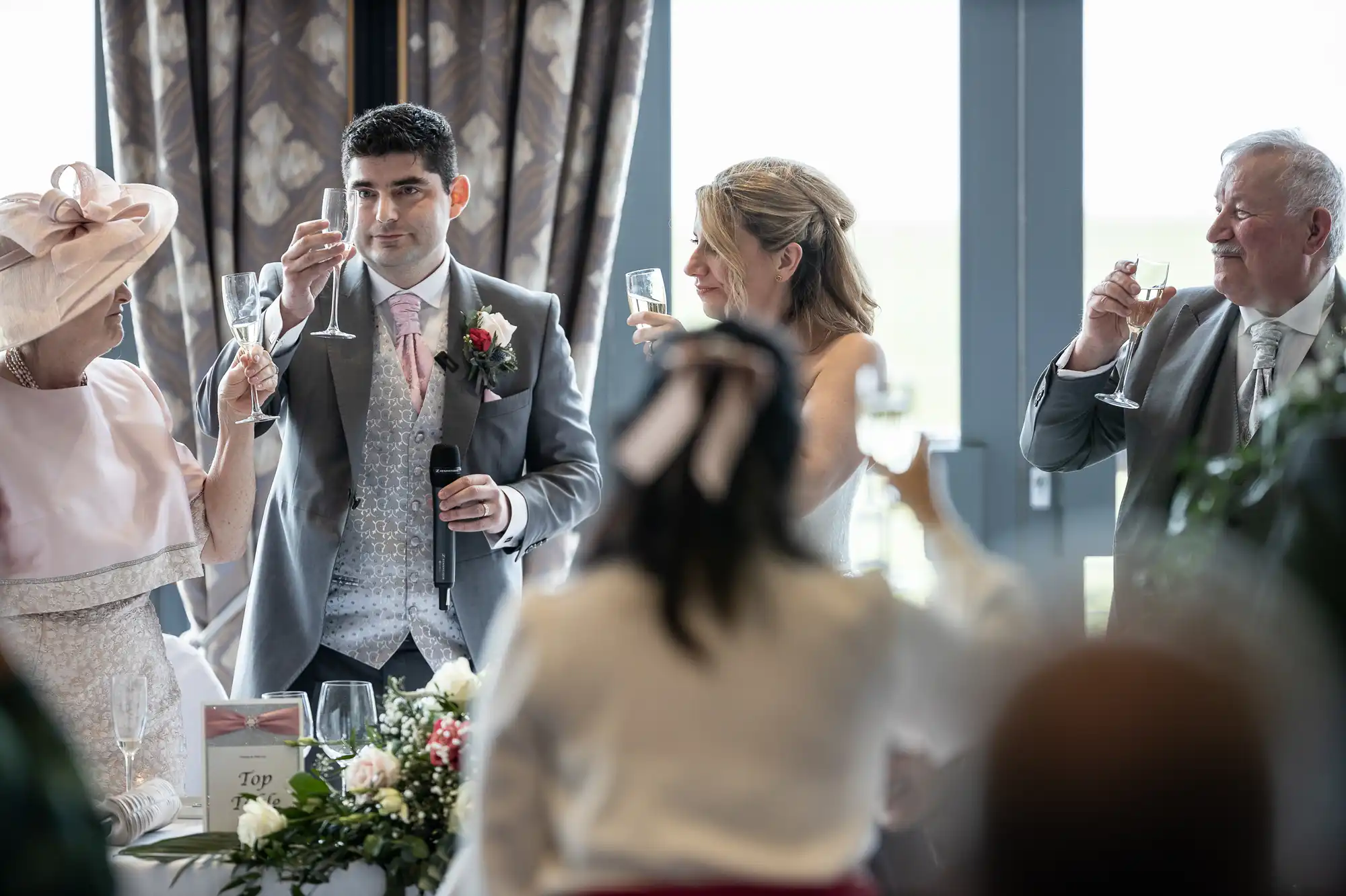 A man in a suit holds a microphone and raises a glass, surrounded by three people also raising glasses in a formal setting. A person in the foreground faces away, wearing a bow on their head.