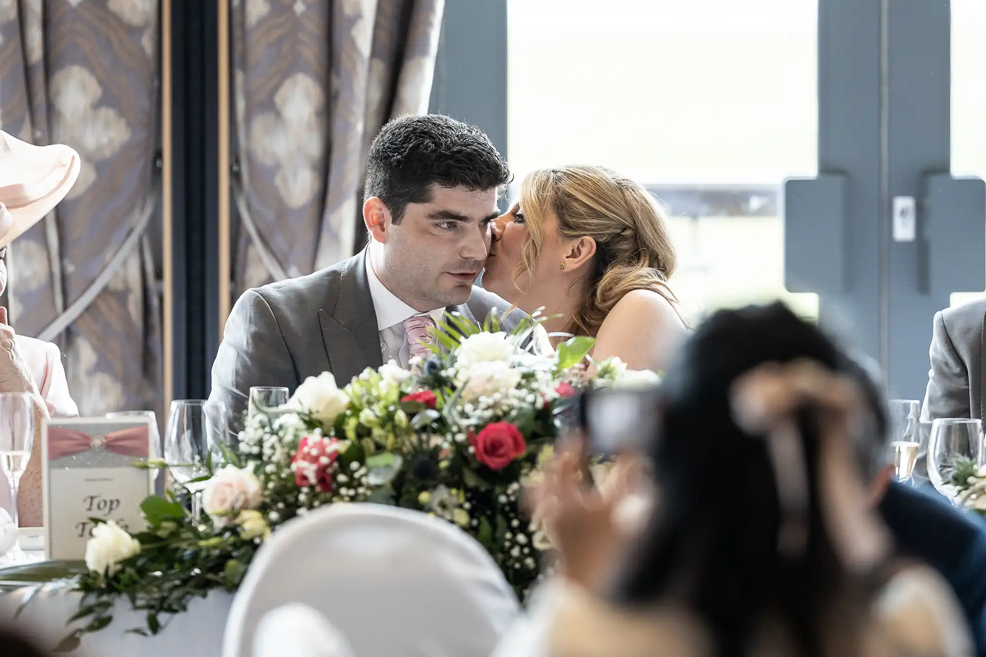 A woman whispers into a man's ear while sitting at a table adorned with flowers and decorations. They are dressed in formal attire in what appears to be a wedding reception setting.