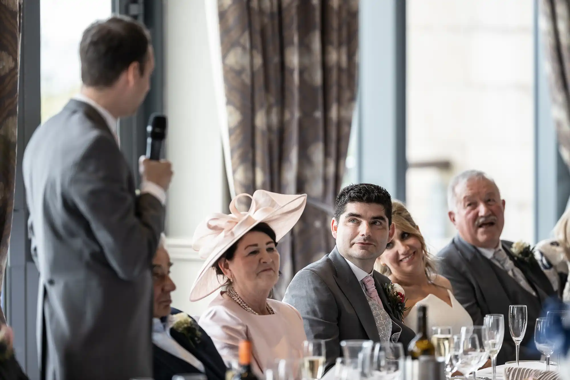 A man in a suit gives a speech with a microphone at a formal event. Four people are seated at a table, listening attentively, with drinks and table settings visible.