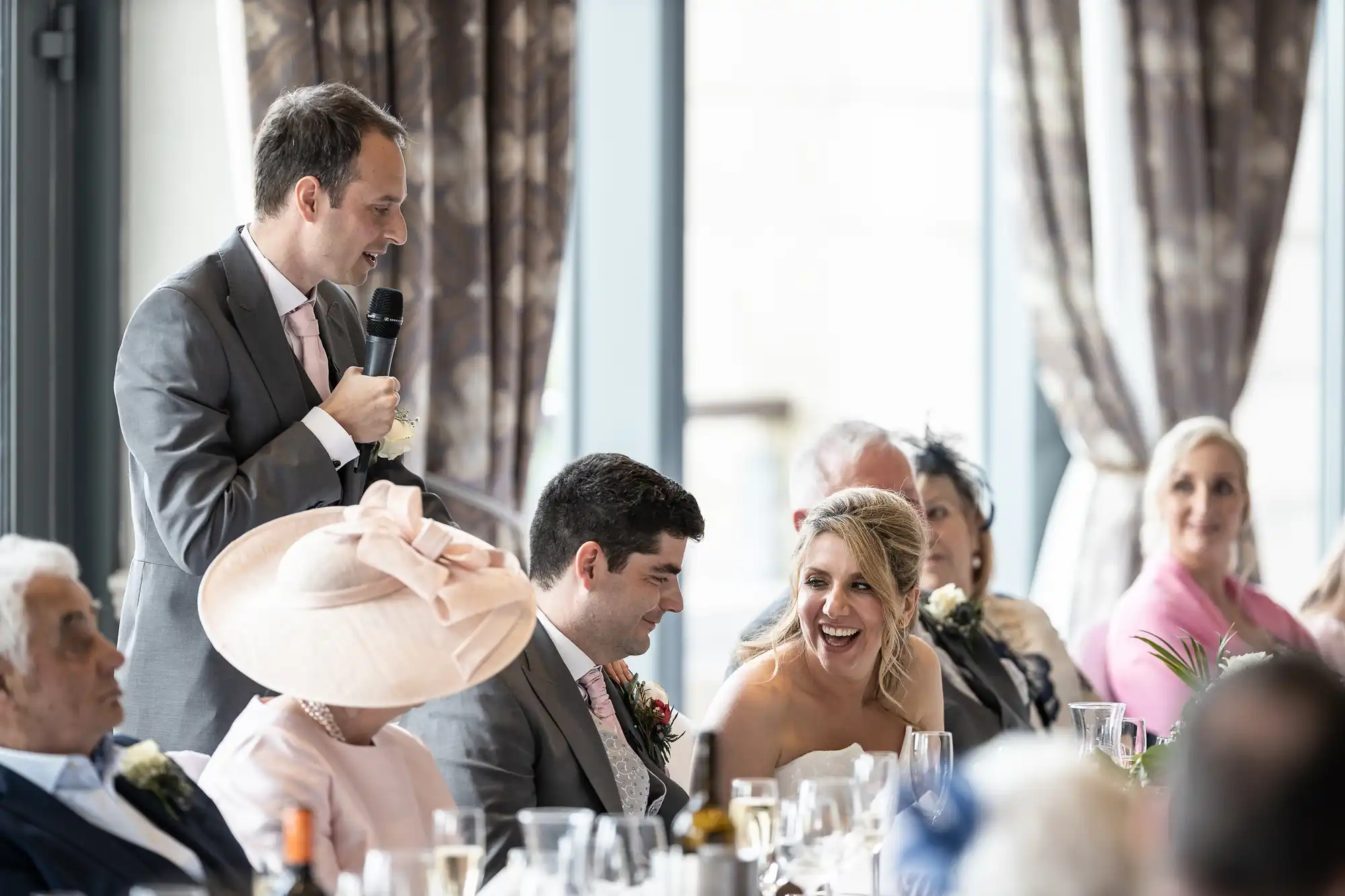 A man in a suit gives a speech while holding a microphone at a wedding reception. The bride and groom are seated and smiling along with other guests around a table.