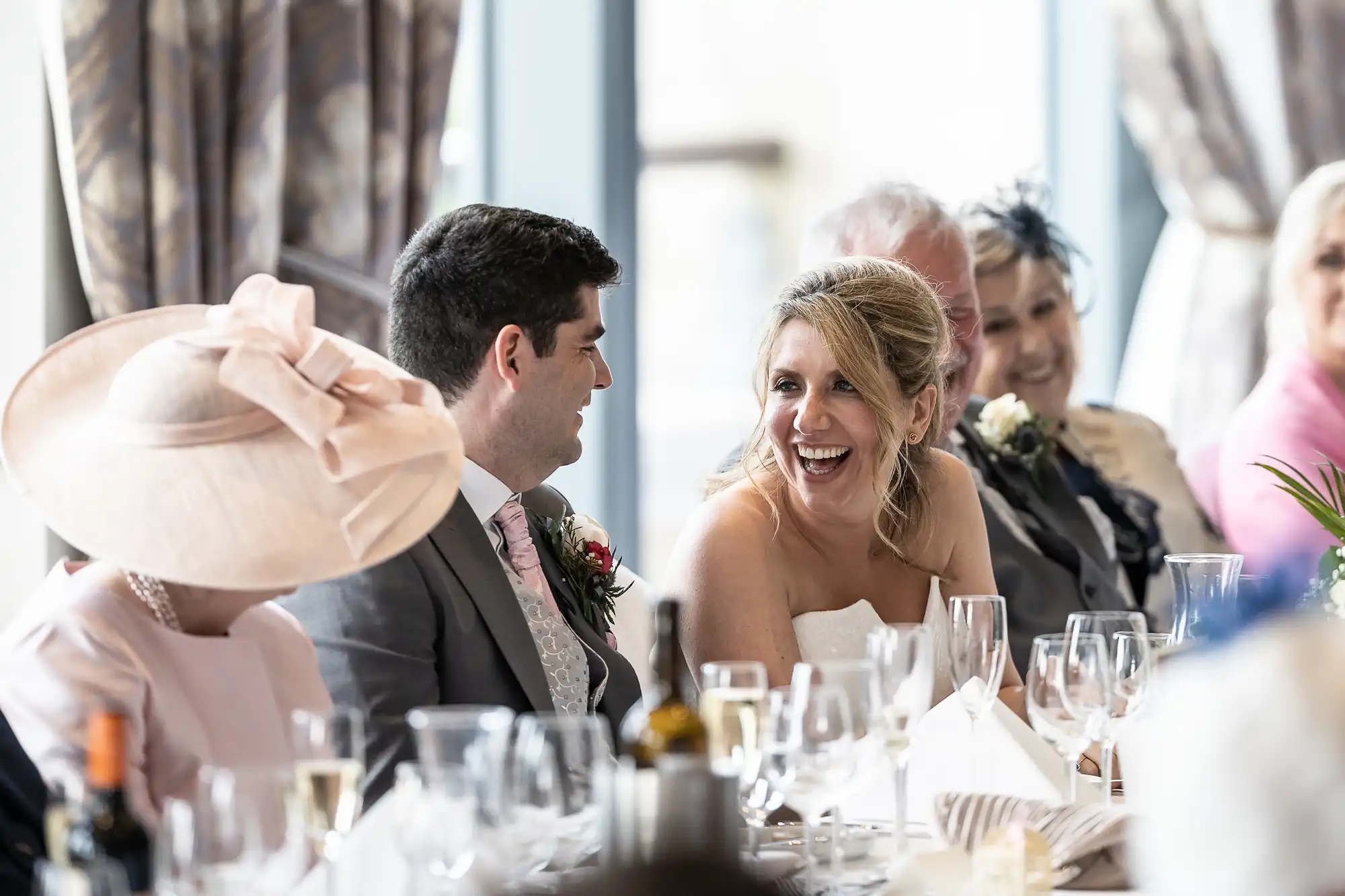 A bride and groom dressed in wedding attire smile and laugh while seated at a long table with other guests during a wedding reception.