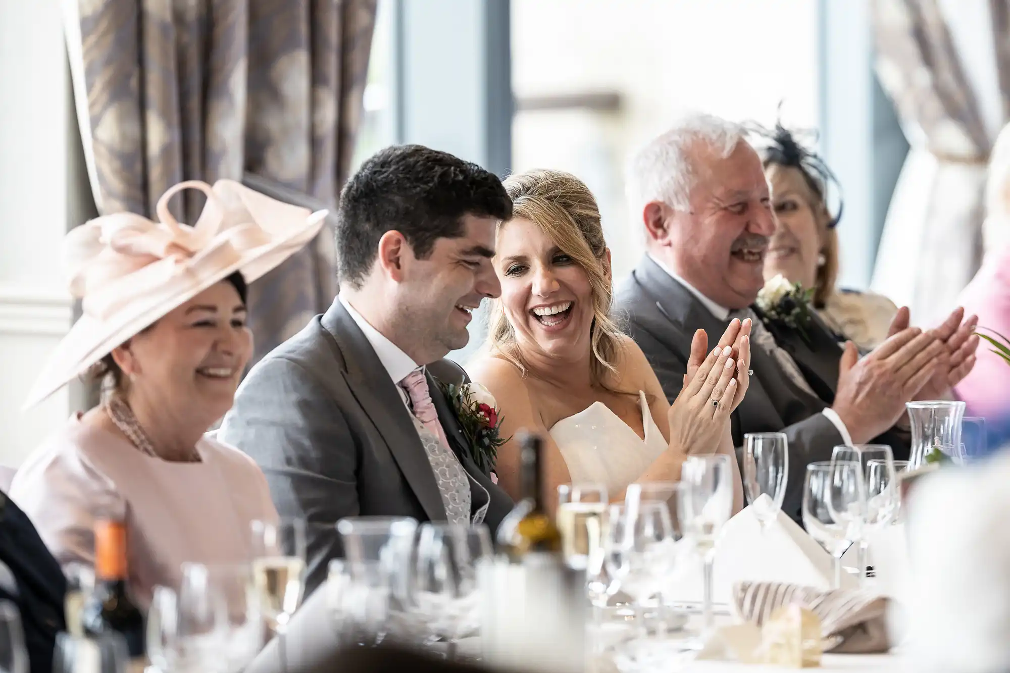 A wedding reception scene with a group of people sitting at a table. They are smiling and clapping, with glasses and table decorations in front of them.