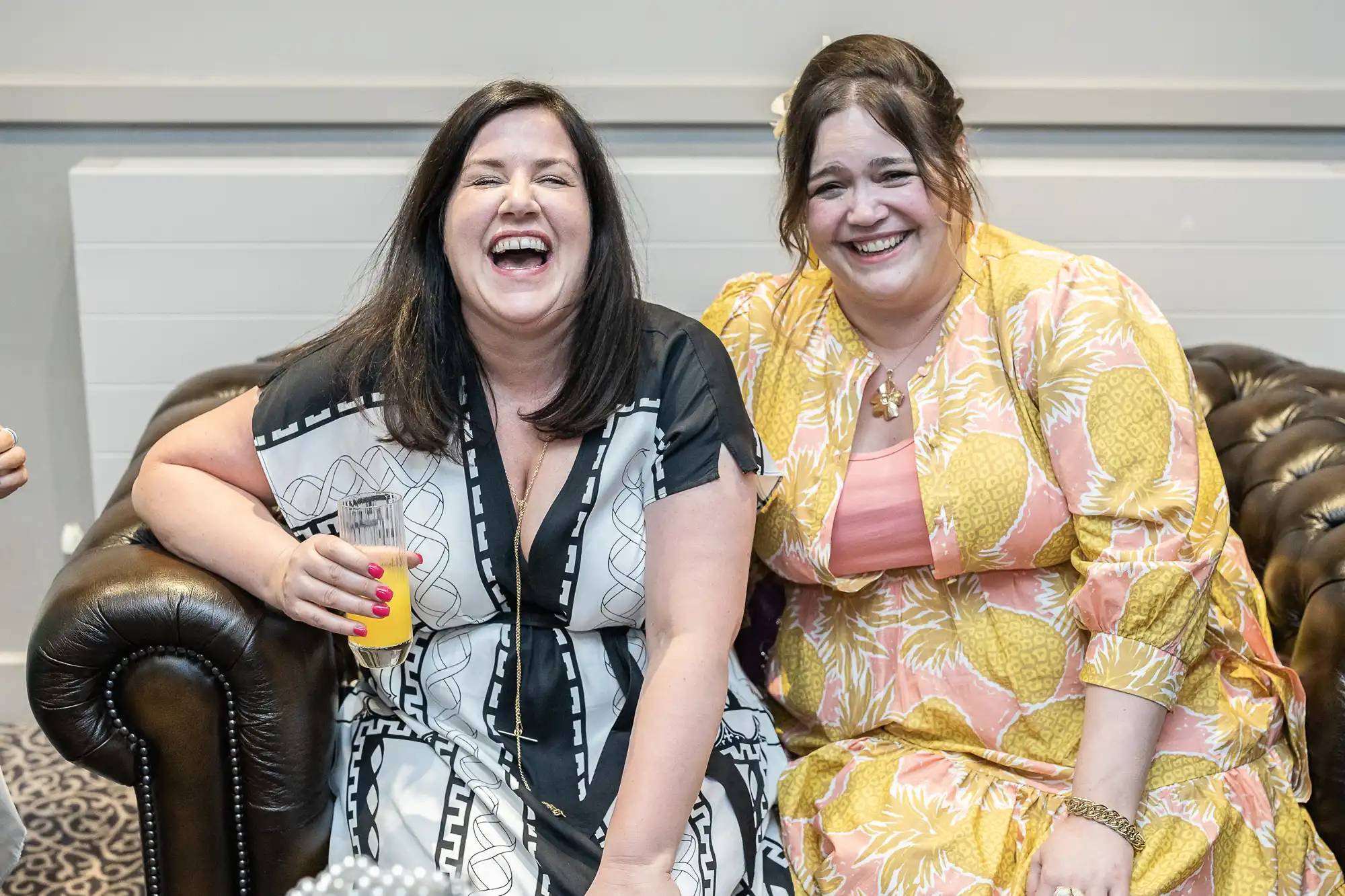 Two women sit on a brown leather couch, smiling and laughing. One holds a drink and wears a black and white dress, while the other is in a yellow patterned dress.