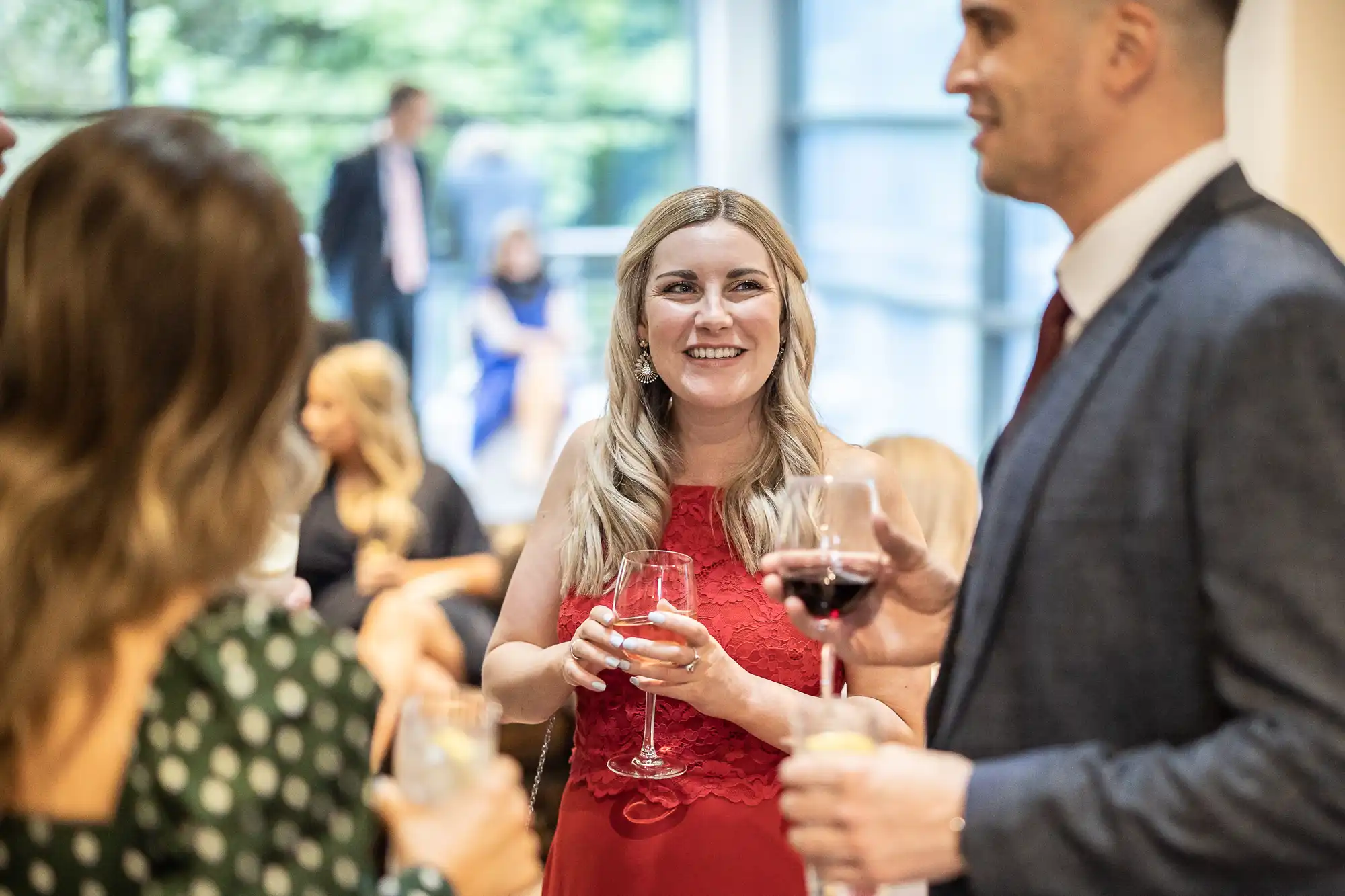 A group of people stand indoors, holding glasses and engaging in conversation. One woman in a red dress is smiling. The background is out of focus, showing more people and greenery through a window.