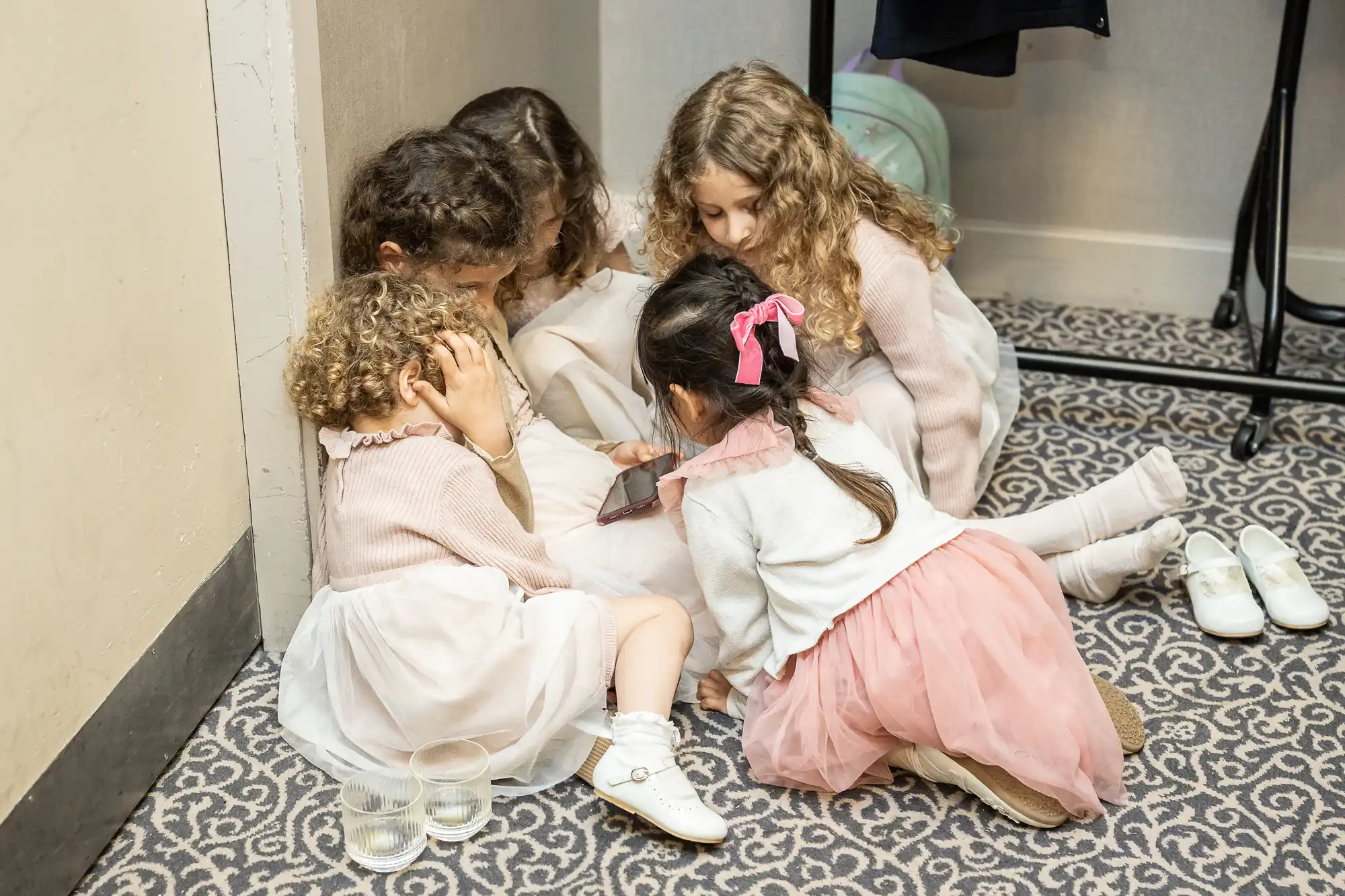 Four young girls wearing pink and white dresses sit closely together on a patterned carpet, intently looking at a smartphone. There are two clear glasses on the floor beside two of the girls.