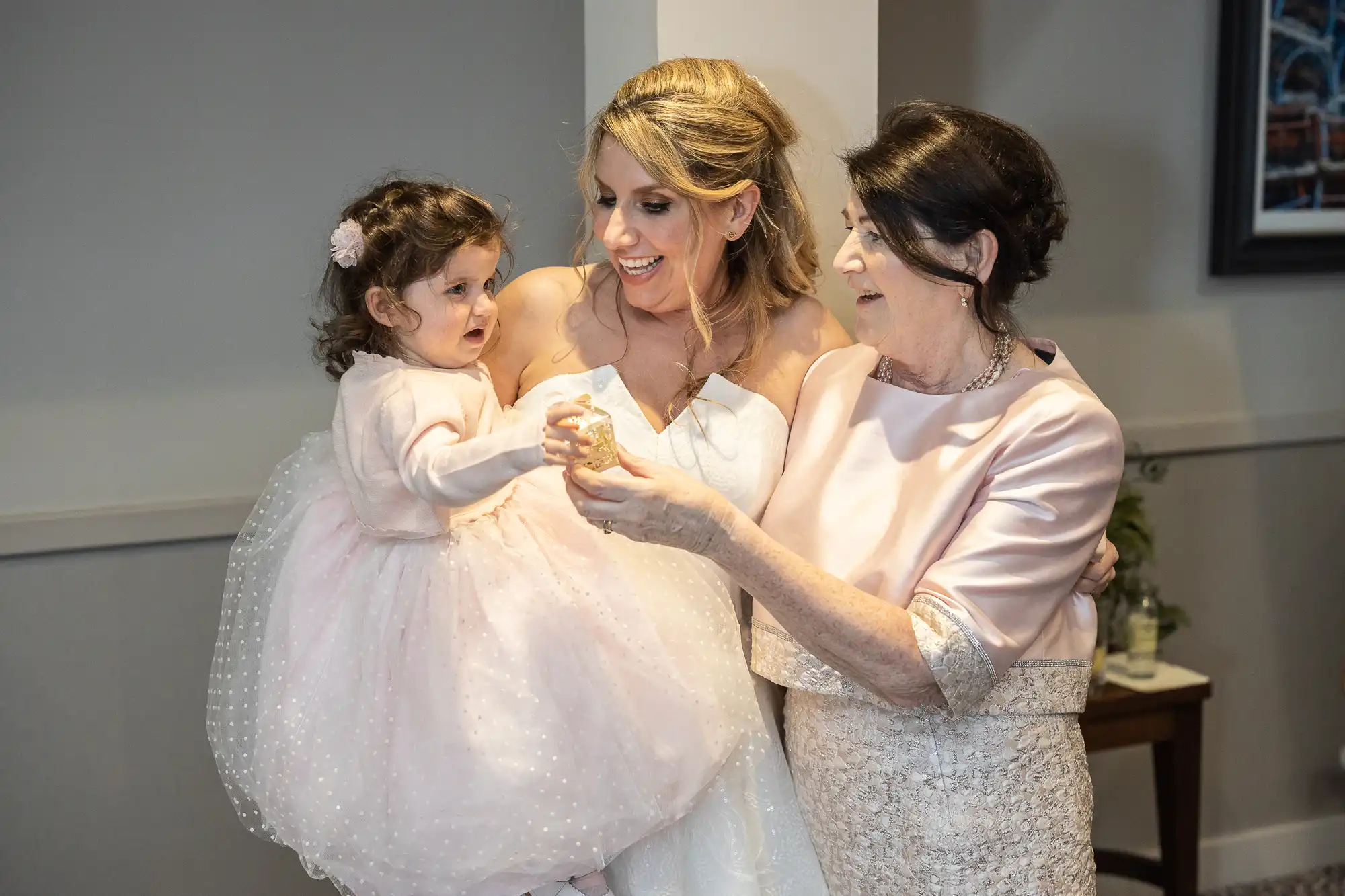 Three generations: A woman in a white dress holds a young girl in a pink dress, standing next to an elderly woman in a pink outfit. They are indoors, smiling and interacting.