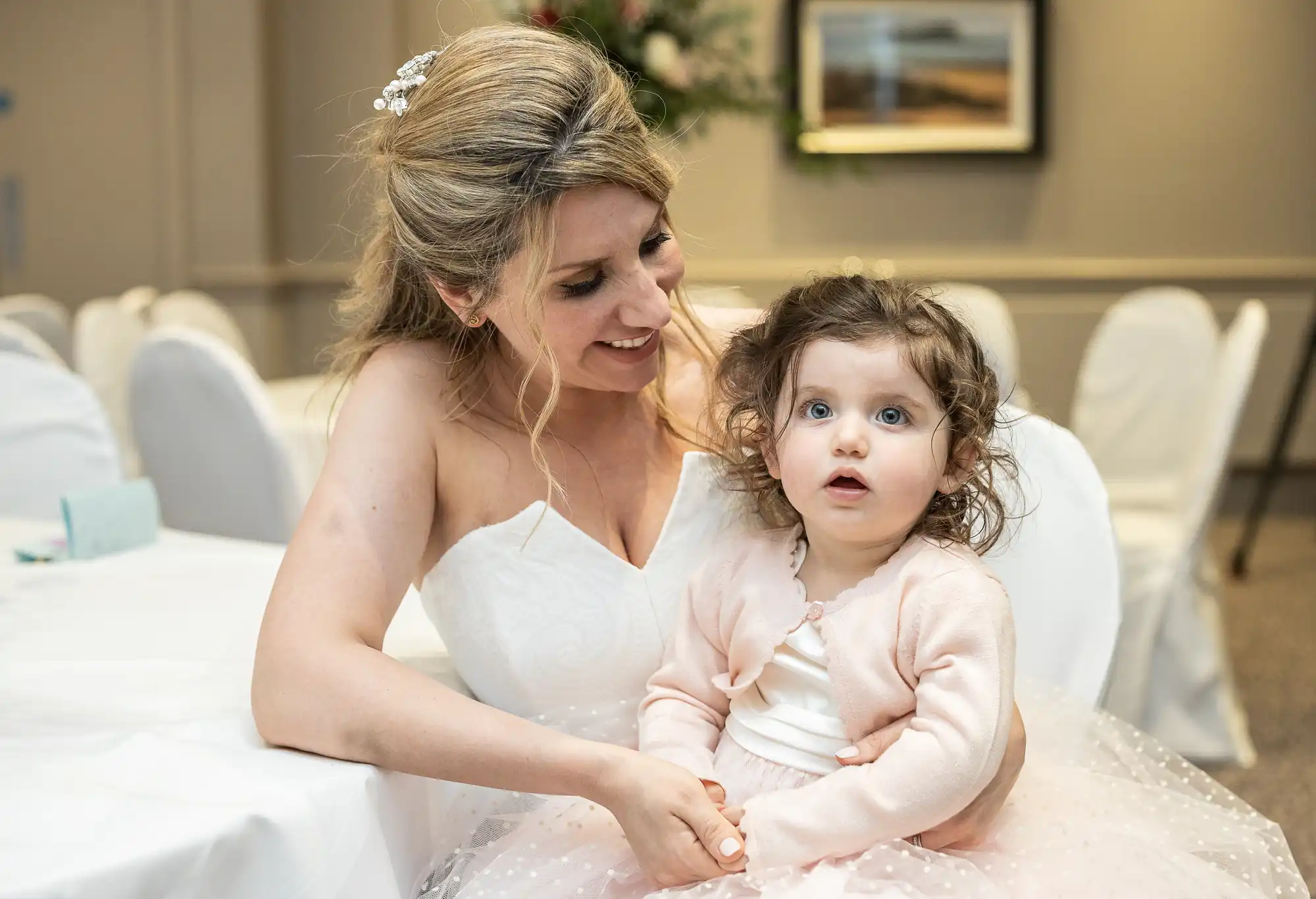 A woman in a white dress smiles while sitting next to a young girl in a pink dress, both seated at a table in a room with white chairs.