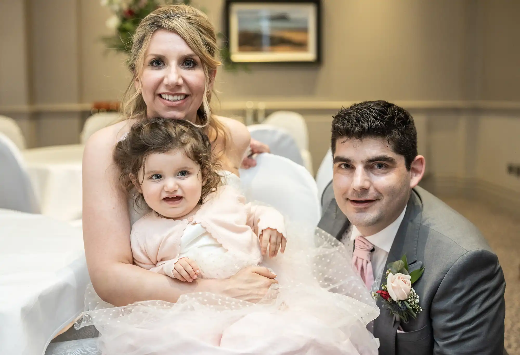 A woman, a man in a suit with a boutonniere, and a toddler in a white dress pose together indoors.