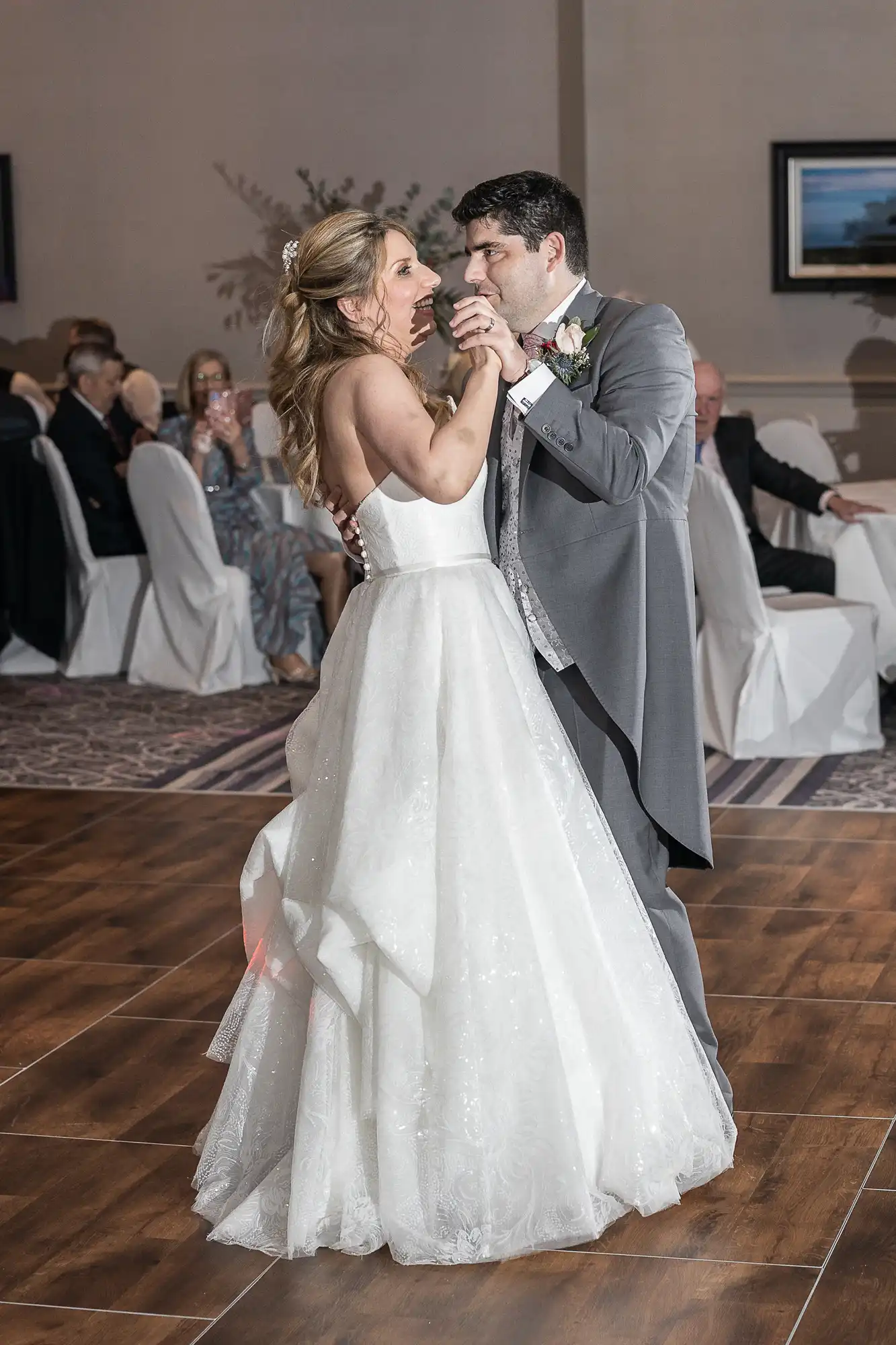 A bride in a white gown and a groom in a grey suit share a first dance at their wedding reception, surrounded by seated guests.