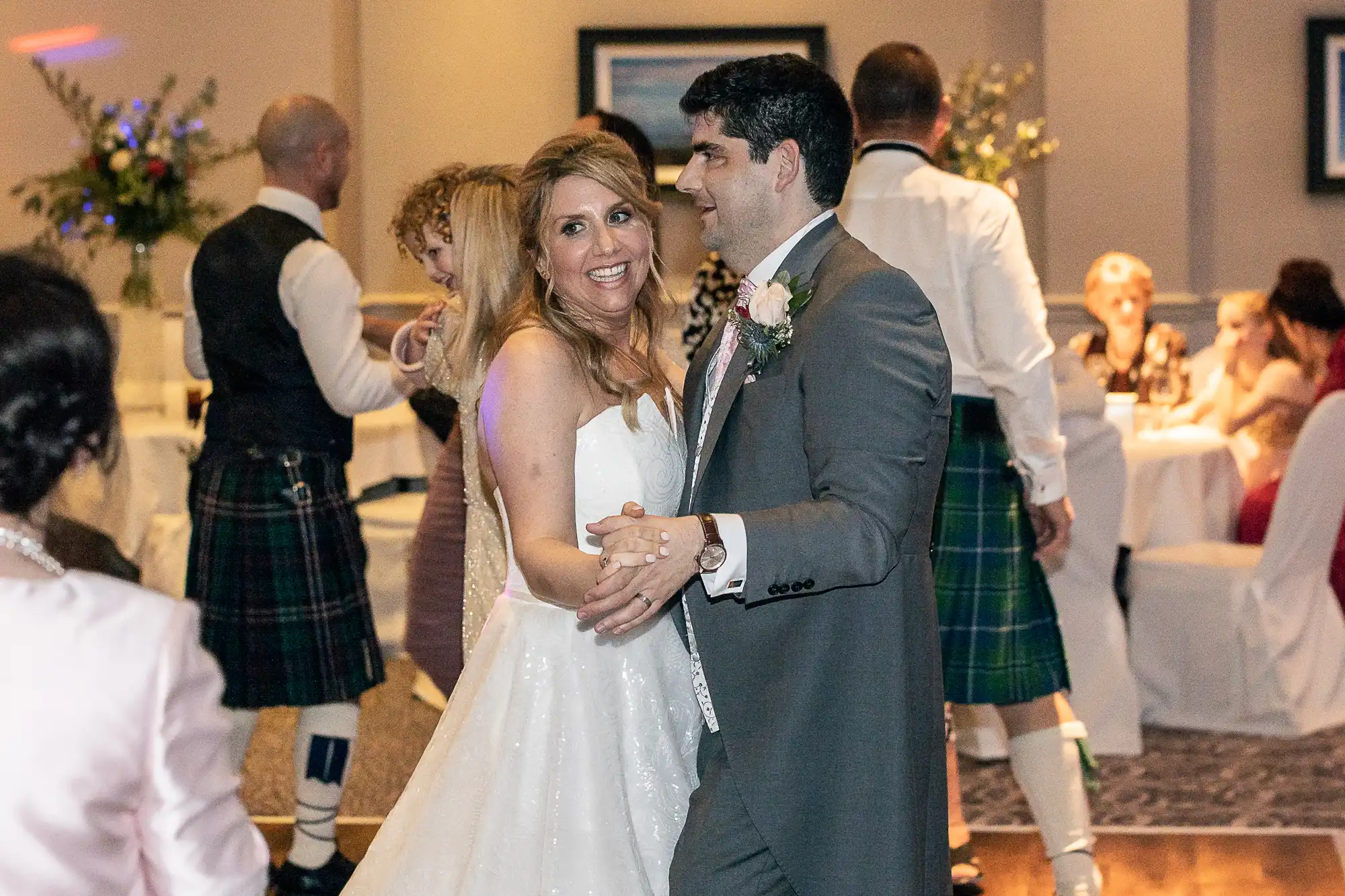 A bride and groom are dancing together at their wedding reception. Guests in formal attire, including some in kilts, are visible in the background.