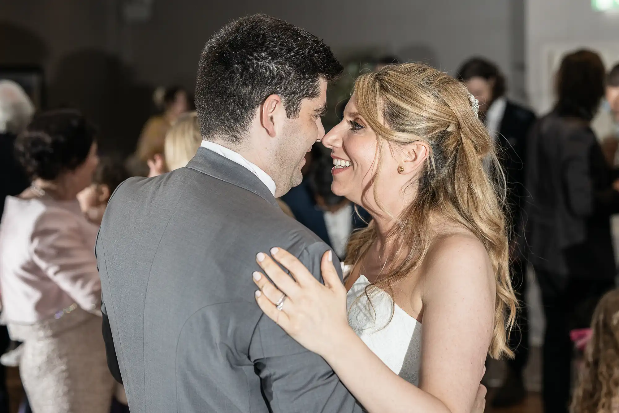 Man in a grey suit and woman in a white dress smiling and dancing together at a social event with other people in the background.