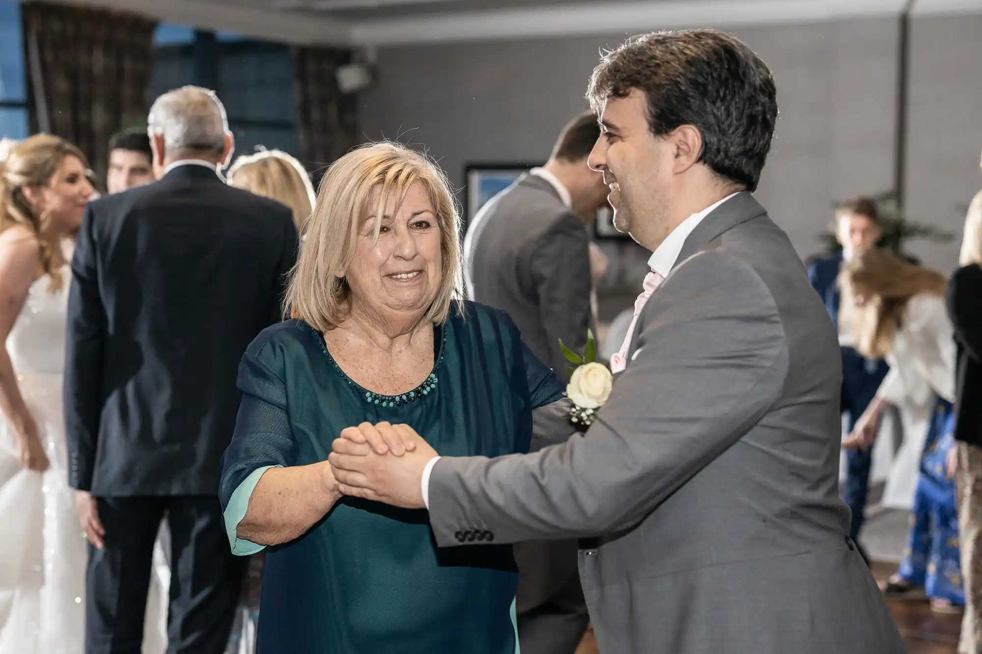 A man in a suit dances with an older woman in a green dress at a formal event, with other attendees visible in the background.