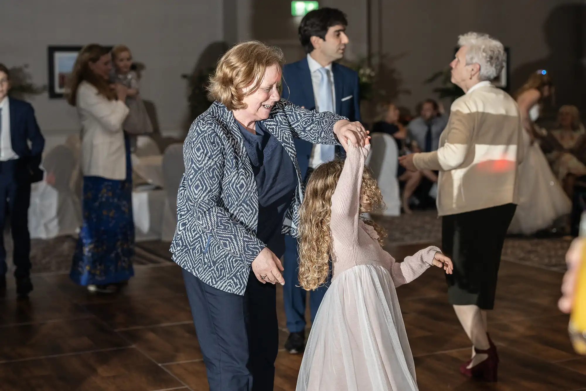 An older woman is dancing with a young girl in a light pink dress at an indoor event. Other attendees are seen in the background, some standing and conversing.