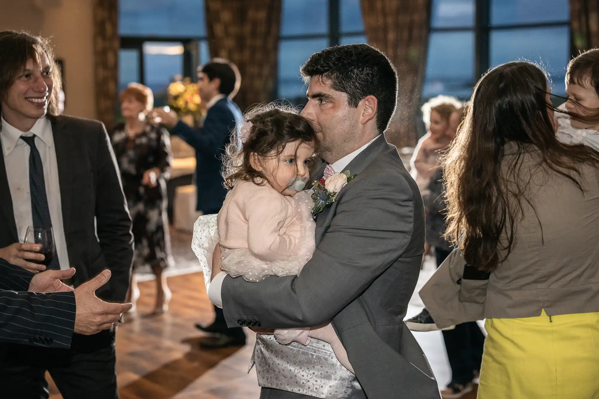 A man in a suit holding a young girl, who is wearing a pale dress, at an indoor social gathering. Several other people are mingling in the background.