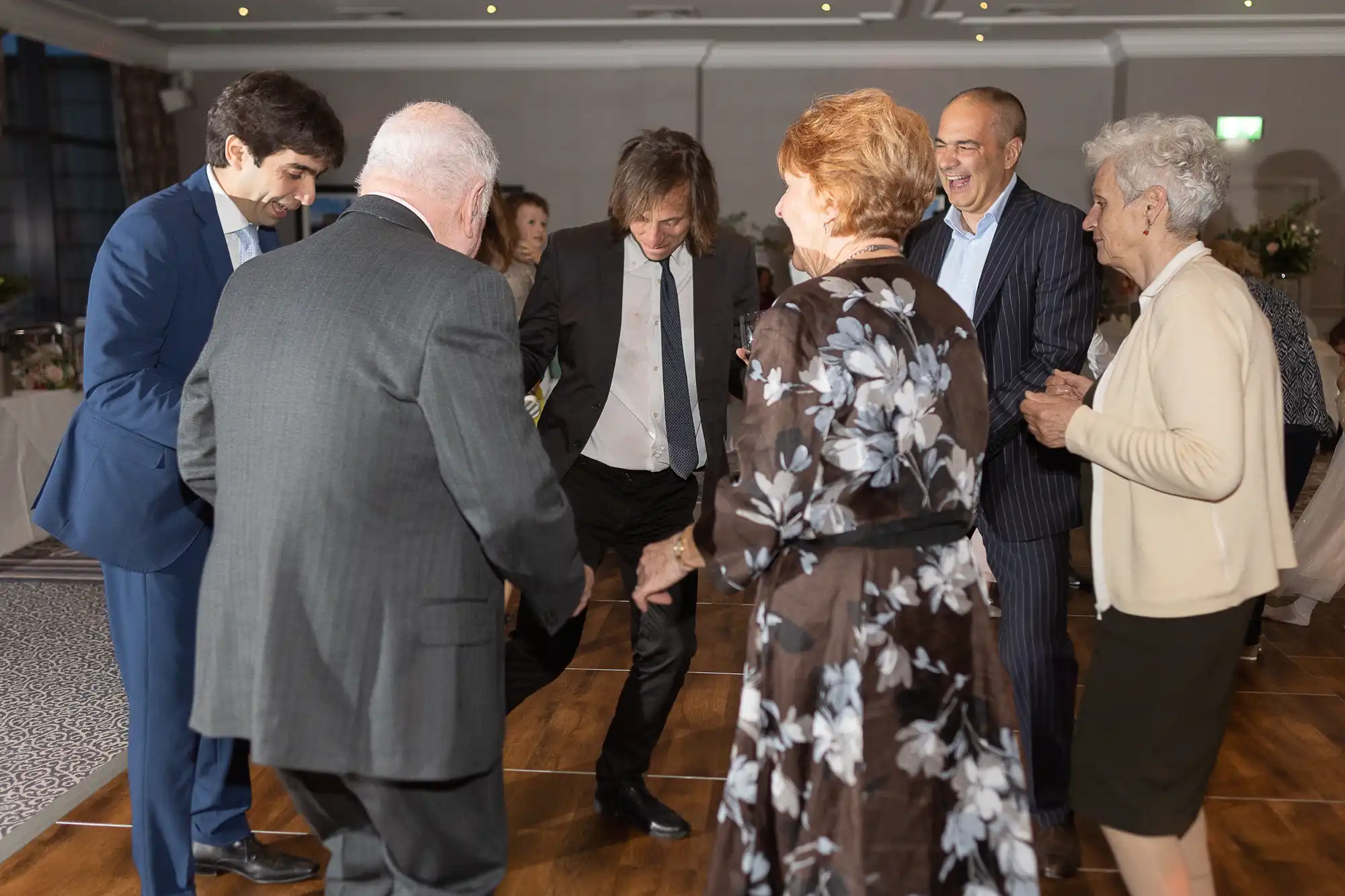 A group of six people, dressed in formal attire, are dancing and smiling together in a well-lit room.