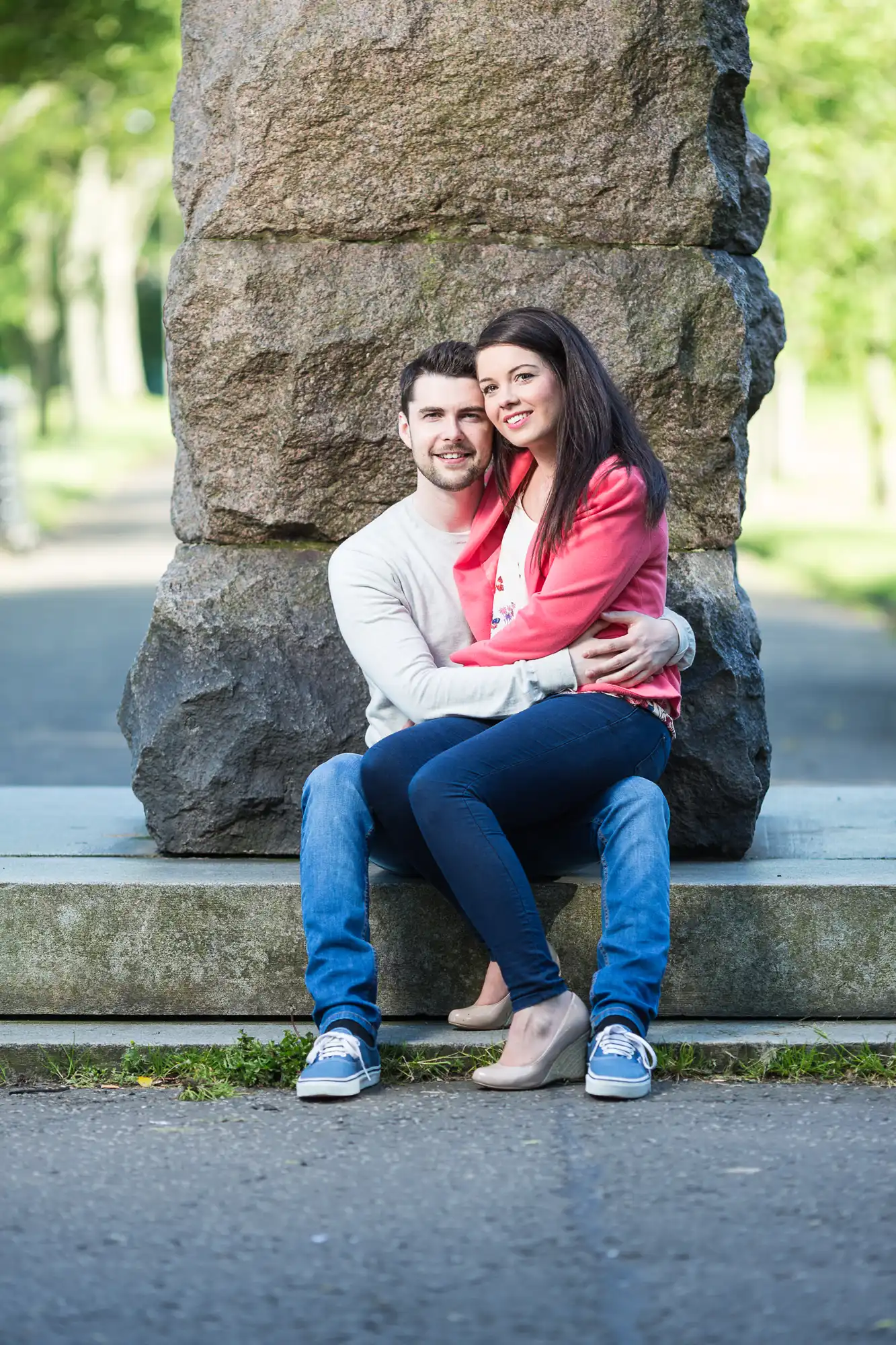 A young couple smiling and sitting closely together on a stone bench in a park, surrounded by trees.