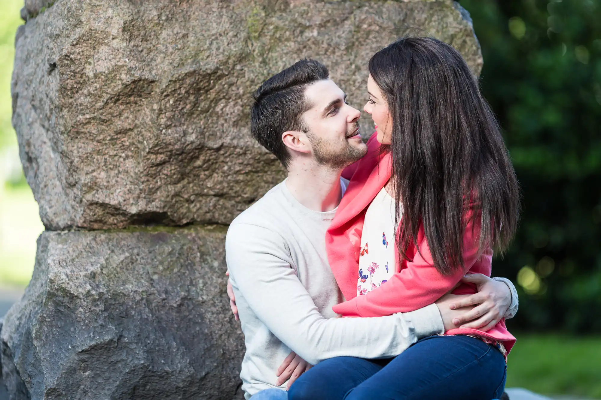 A couple embracing and looking at each other affectionately, seated in front of a large rock in a park.