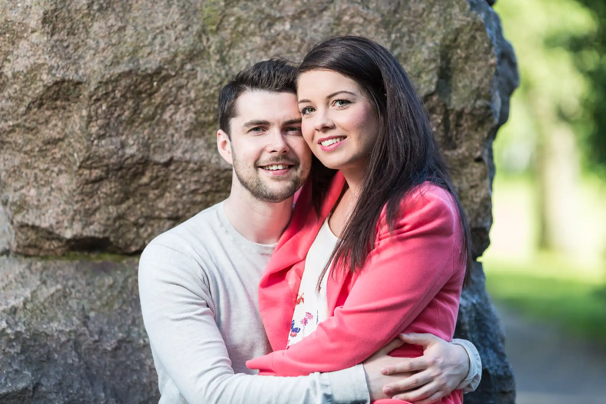 A smiling couple embracing each other in front of a large rock in a sunny park setting.