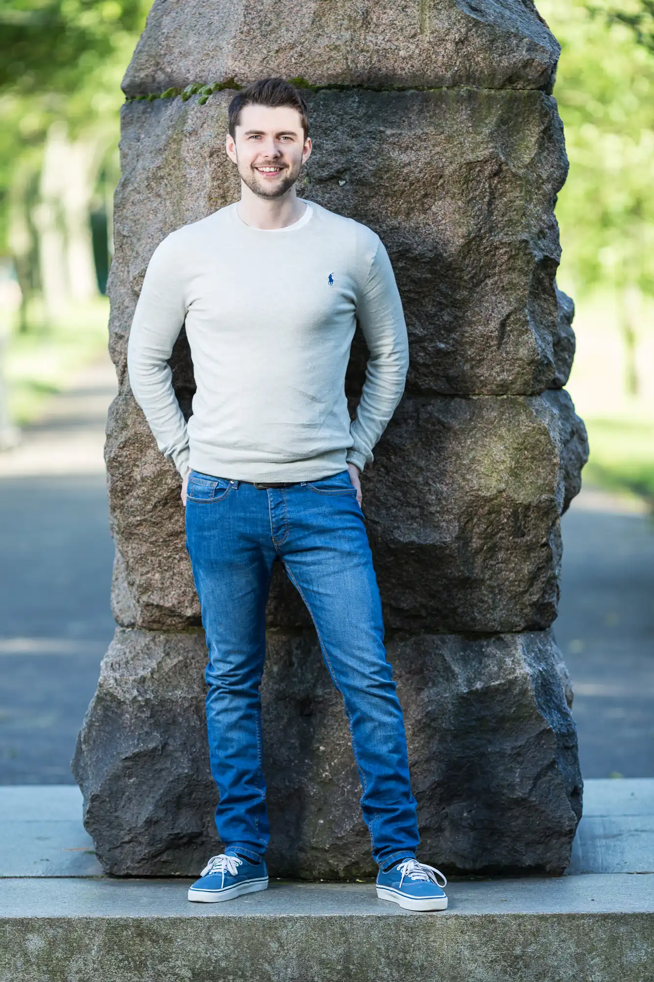 A man smiling for the camera, standing in front of a large boulder in a park, dressed in a white long-sleeve shirt, blue jeans, and blue sneakers.