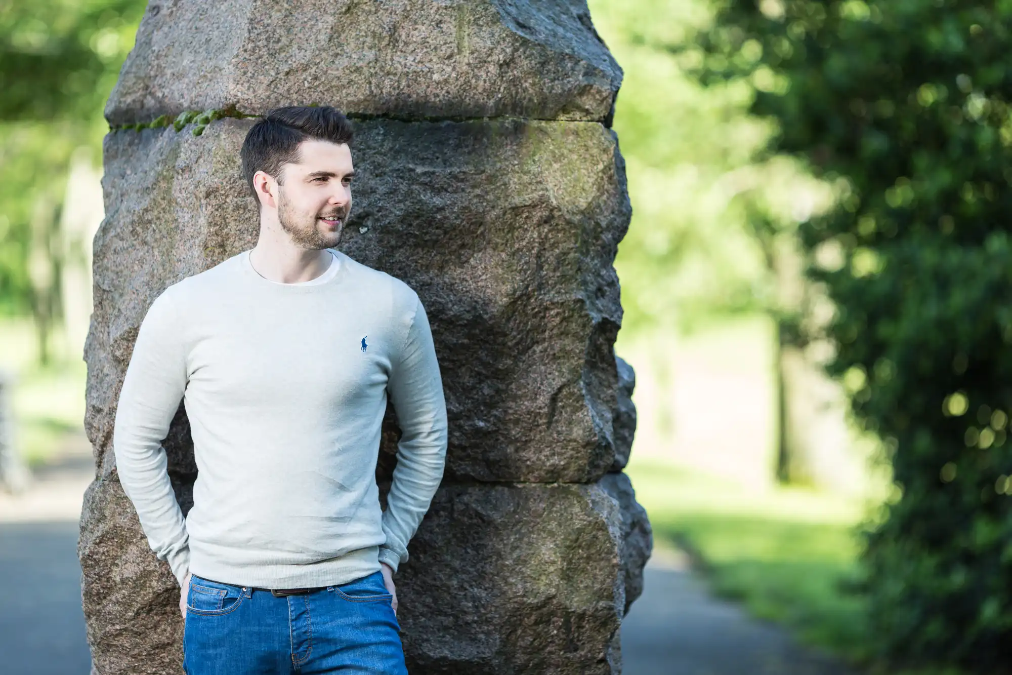 A man in a casual long-sleeve shirt leaning against a large rock in a sunny park, looking to his left with a slight smile.
