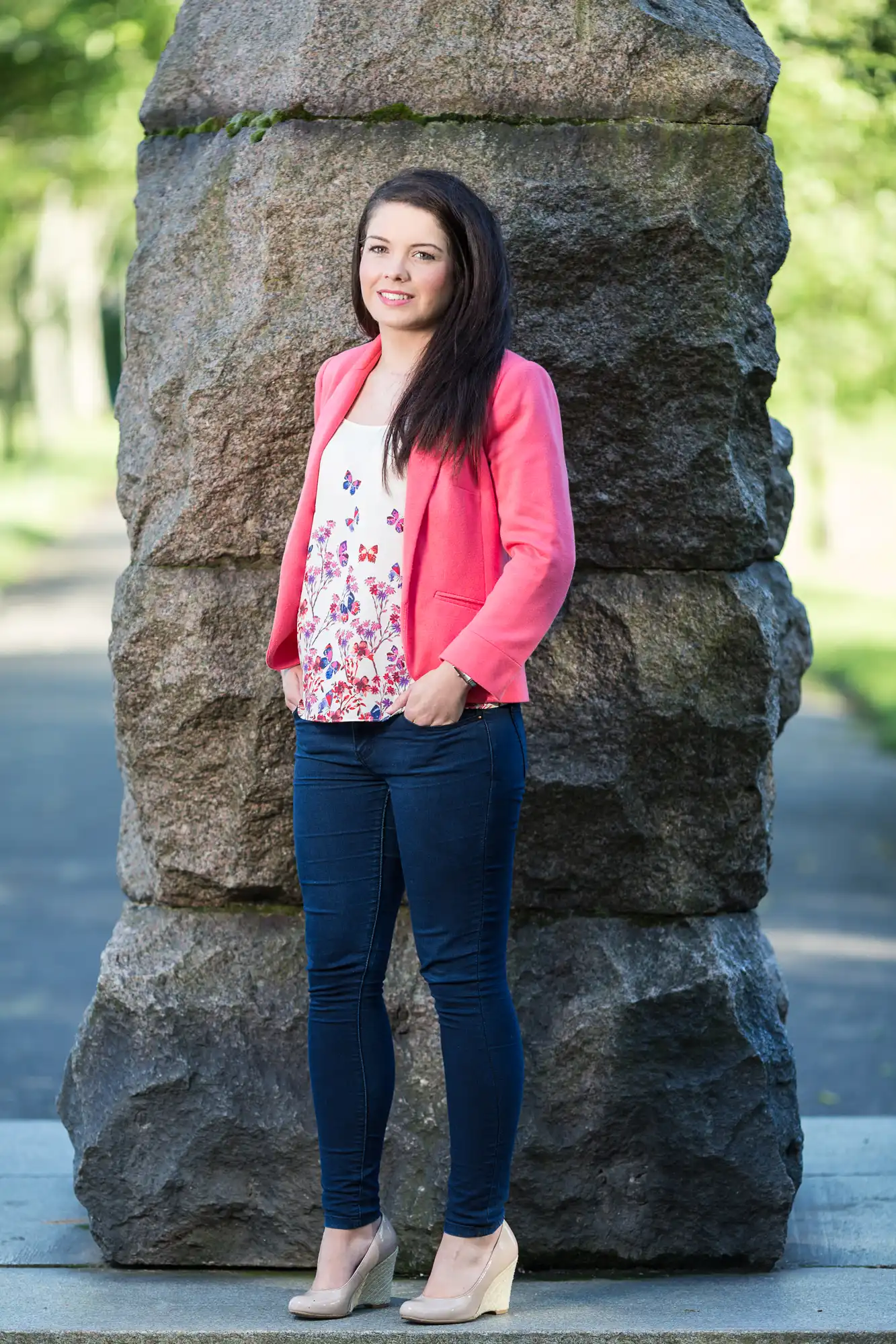 A woman in a pink blazer and blue jeans stands smiling in front of a large stone monument in a park.