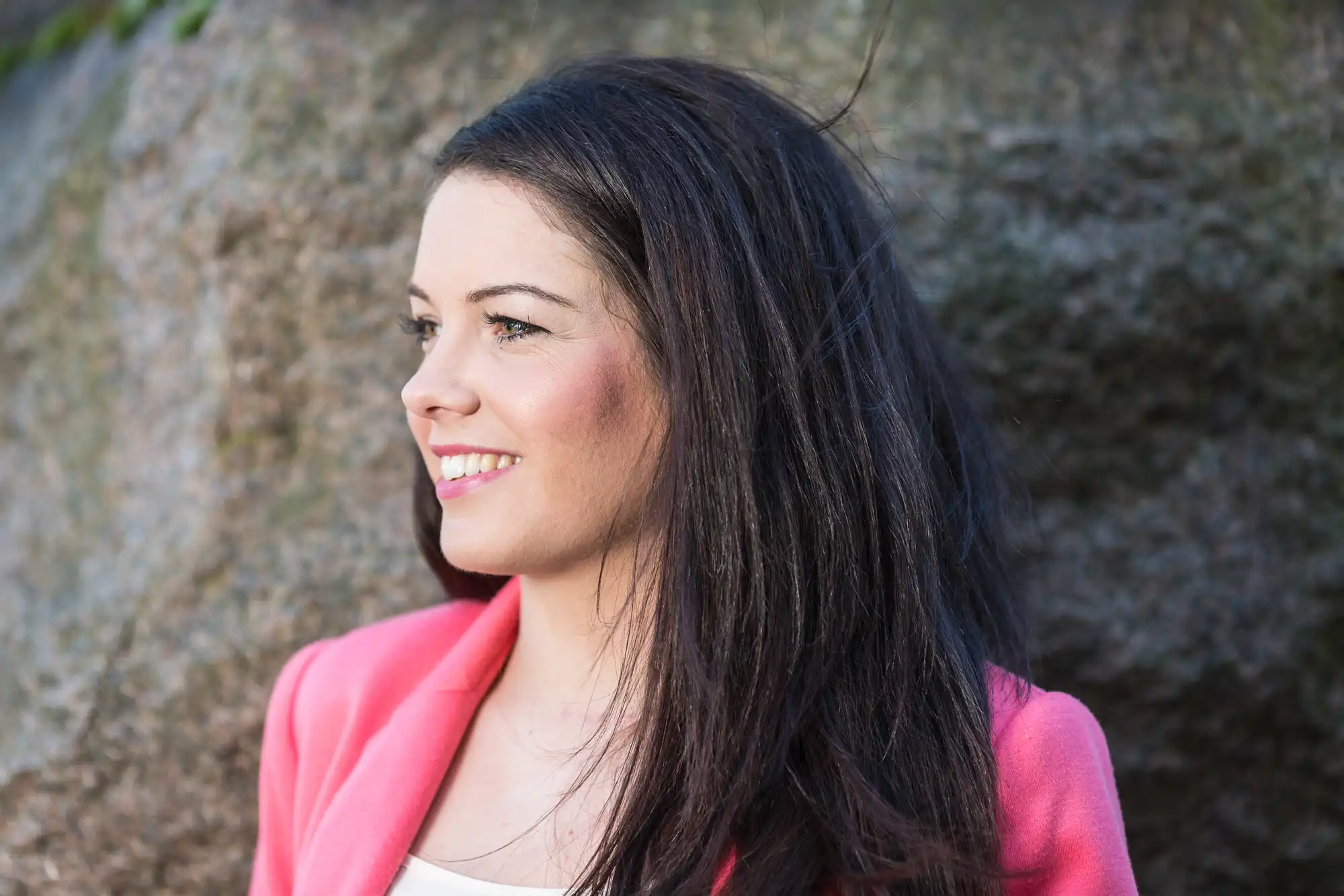 A woman with dark hair wearing a pink blazer smiles while looking to the side, with a large rock in the background.