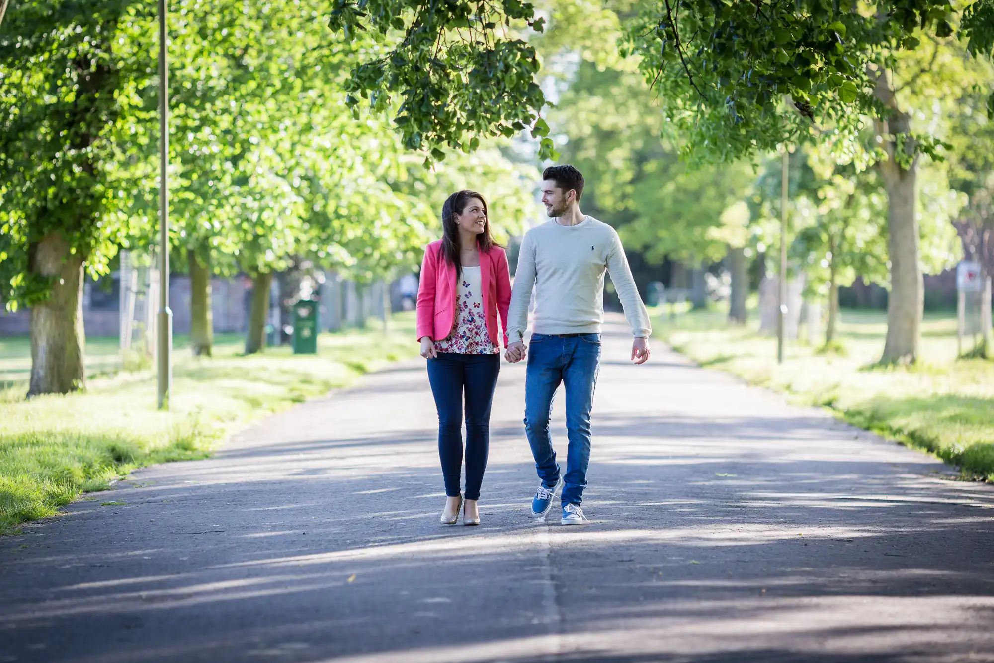 A man and a woman walking and talking on a tree-lined path in a park.