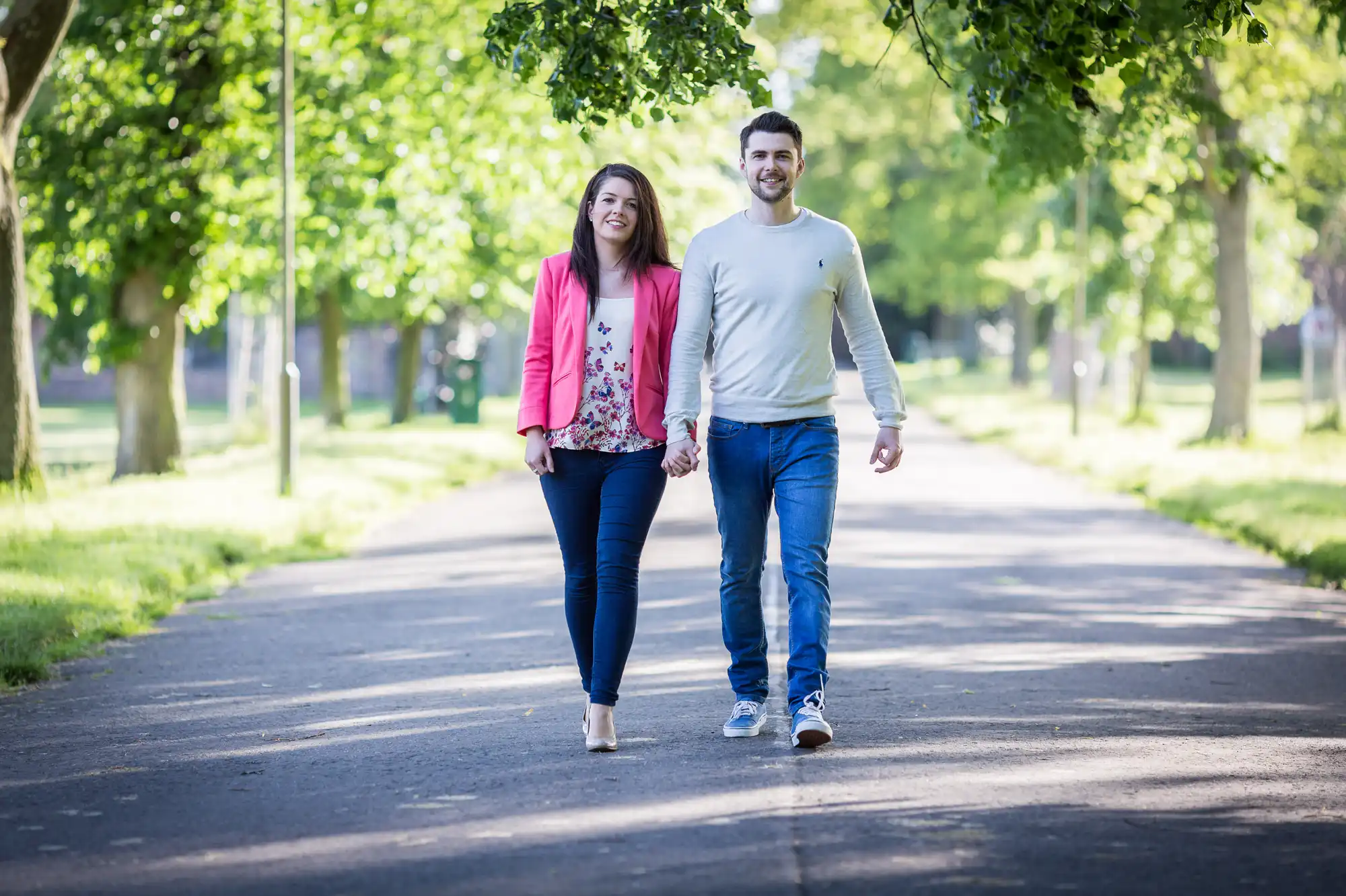 A smiling man and woman holding hands while walking down a tree-lined path on a sunny day.