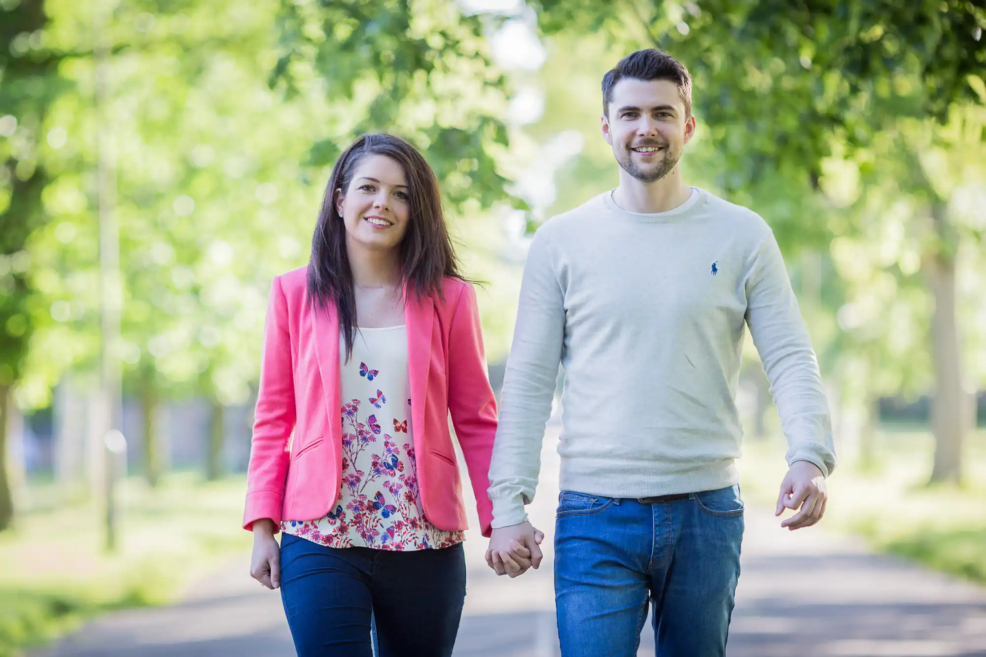 A young man and woman holding hands and smiling while walking down a tree-lined path.