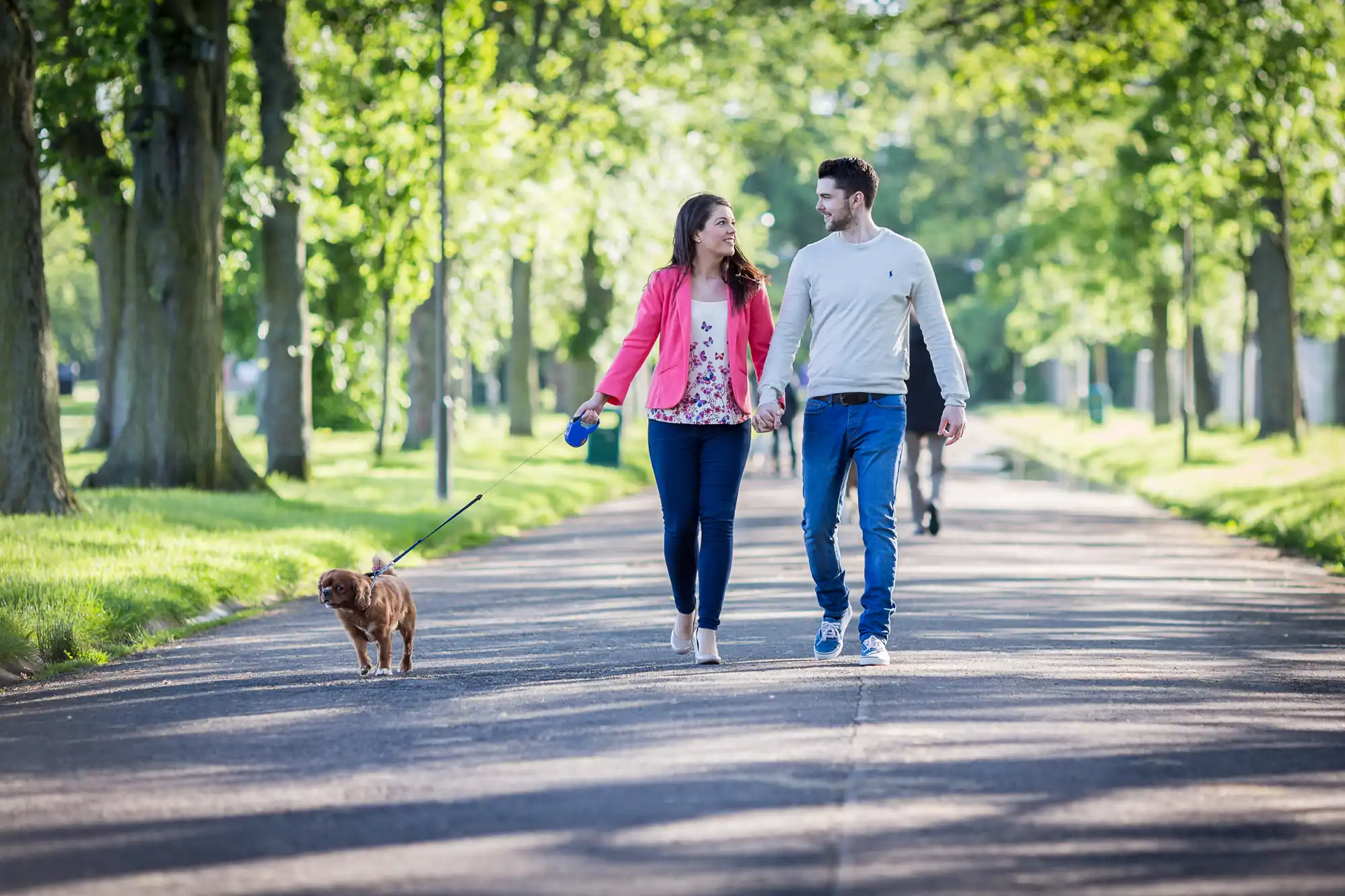 A couple walks hand in hand with a small dog on a leash along a tree-lined park path on a sunny day.