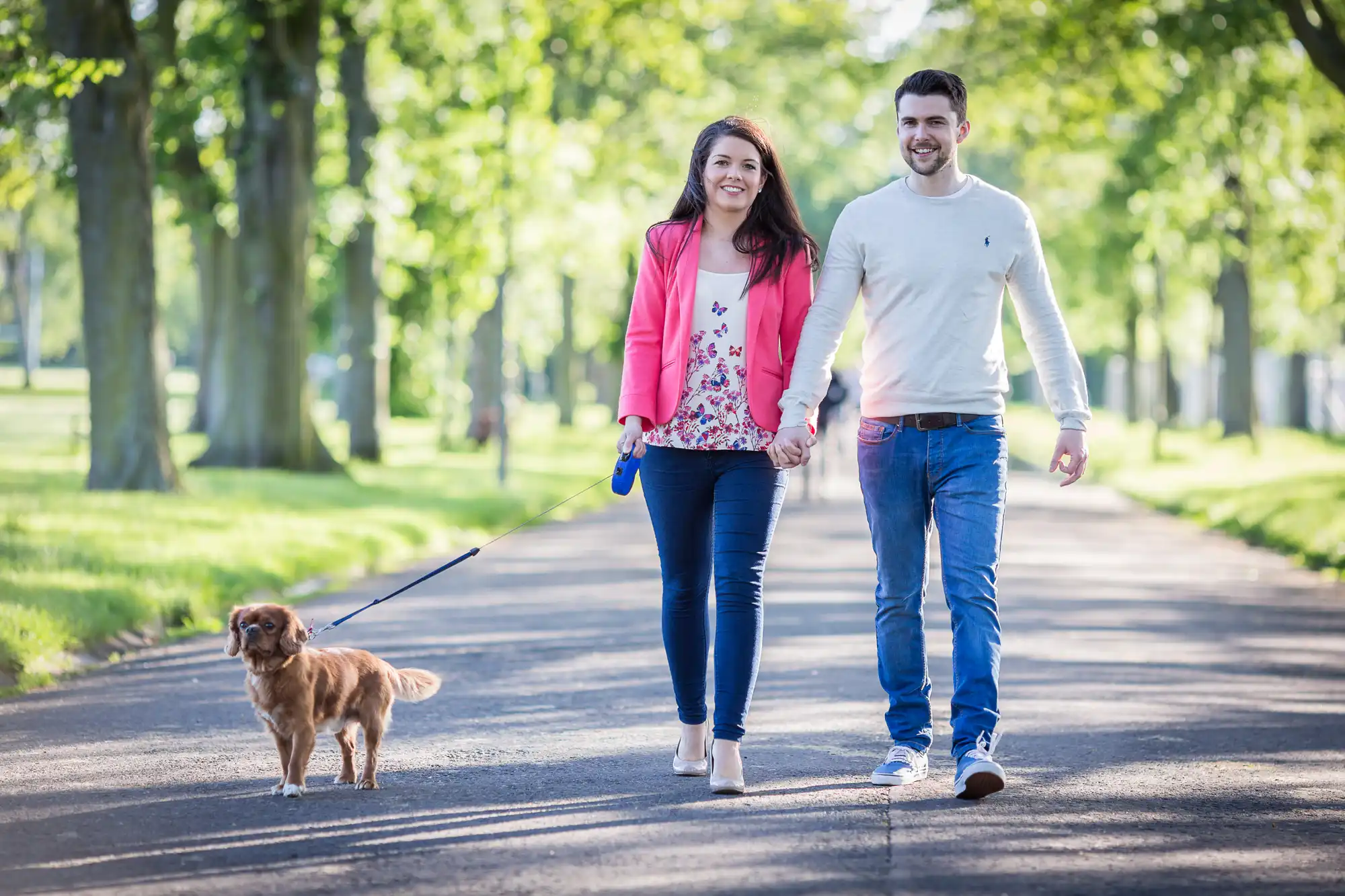 A man and a woman smiling while walking a small brown dog on a leash along a tree-lined pathway.
