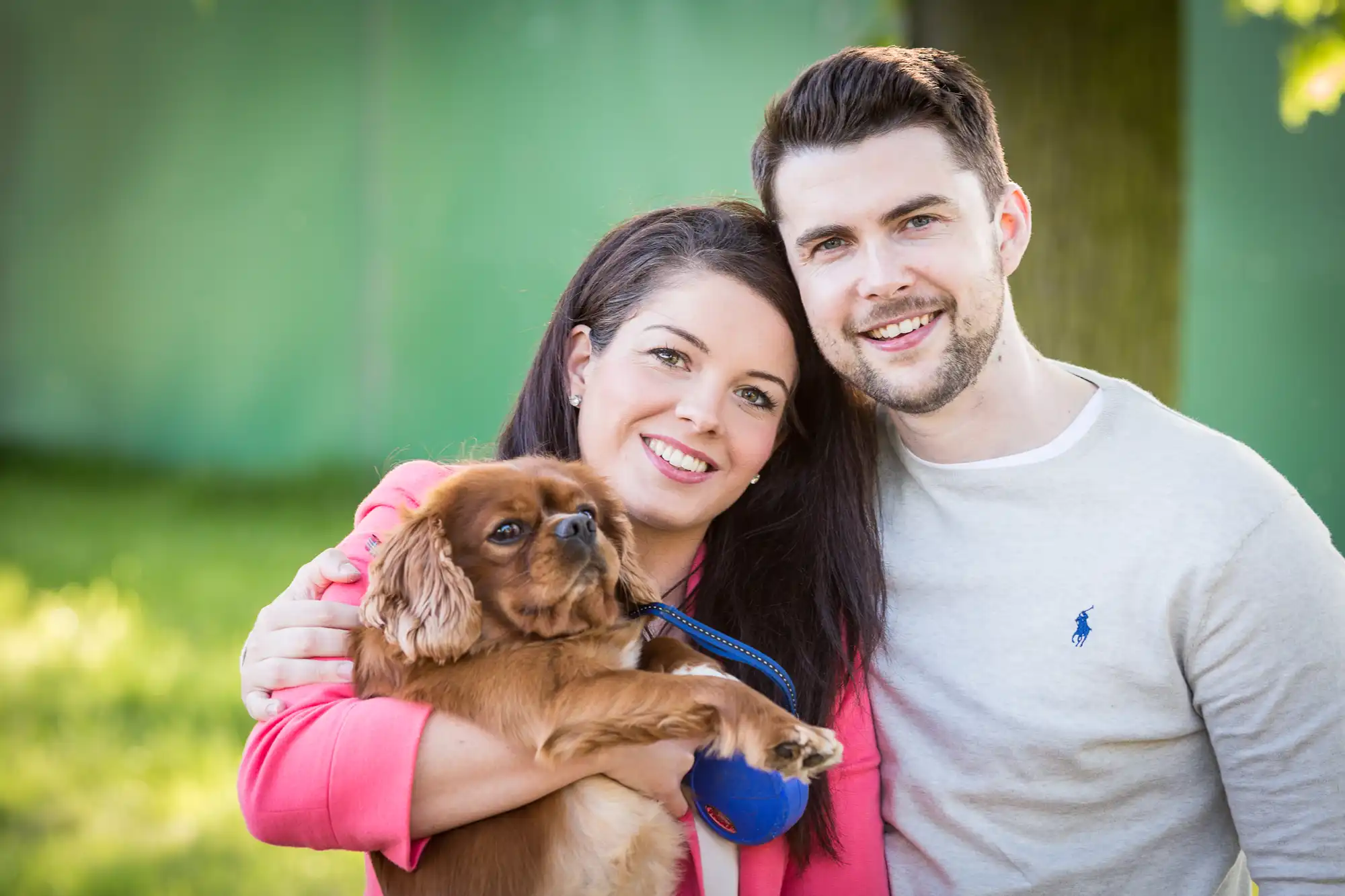 A young couple smiling, holding a small brown dog, standing outdoors in a sunlit park.