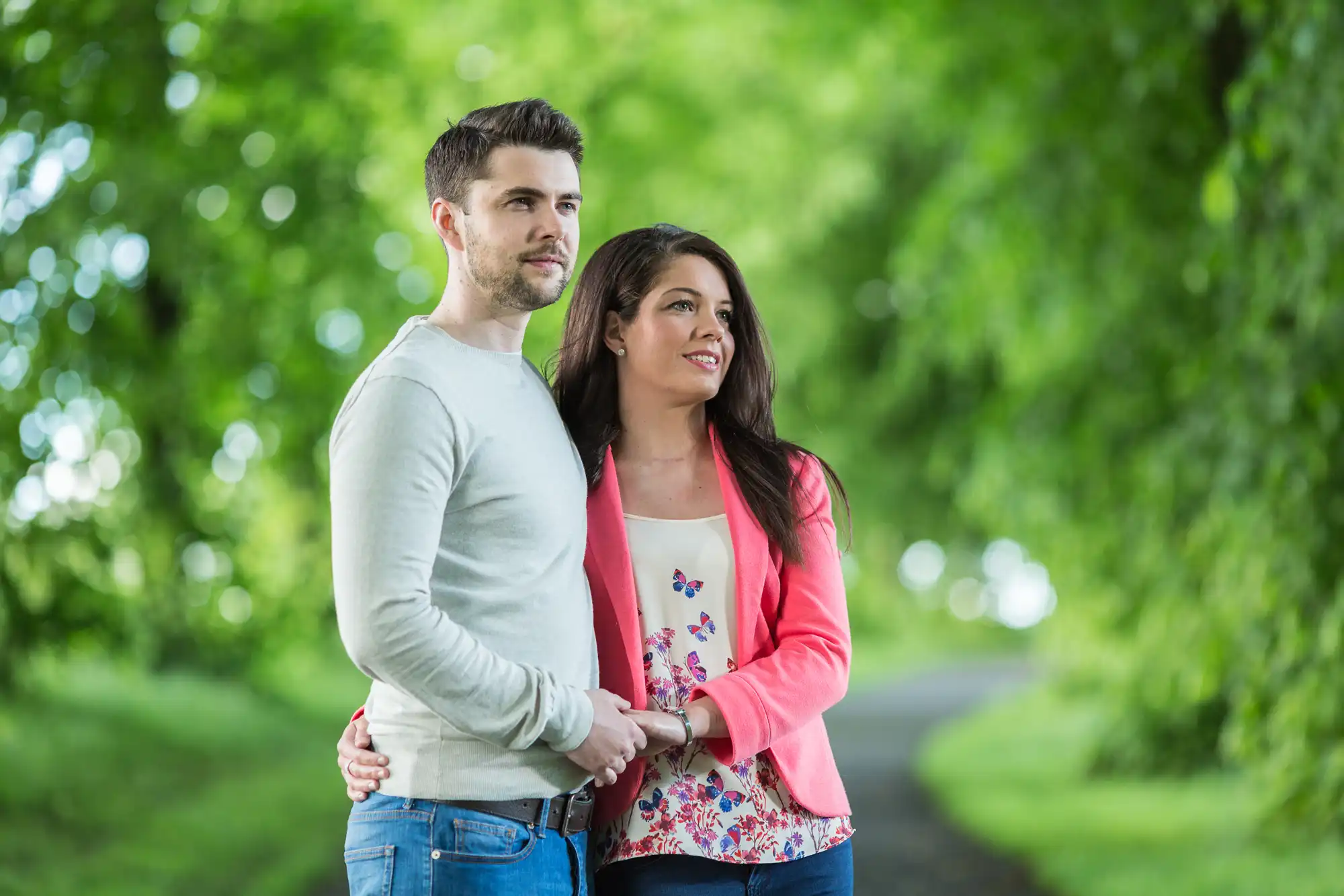 A couple embracing each other, standing on a leafy path in a park, smiling and looking away from the camera.