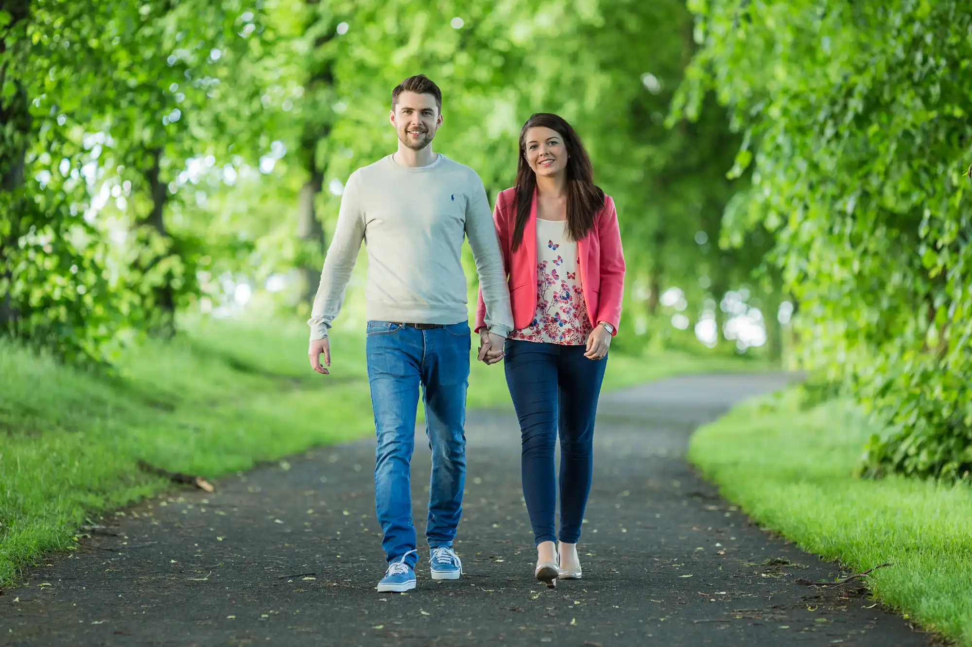 A young couple happily walking hand in hand along a tree-lined path in a green park.