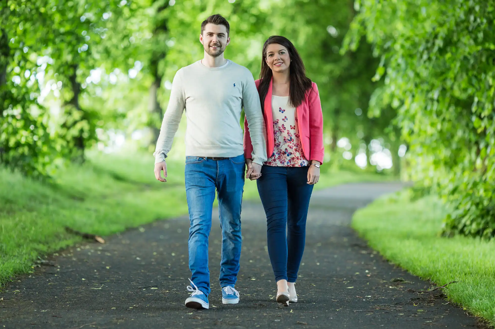 A young man and woman walk hand-in-hand along a tree-lined path, smiling directly at the camera.