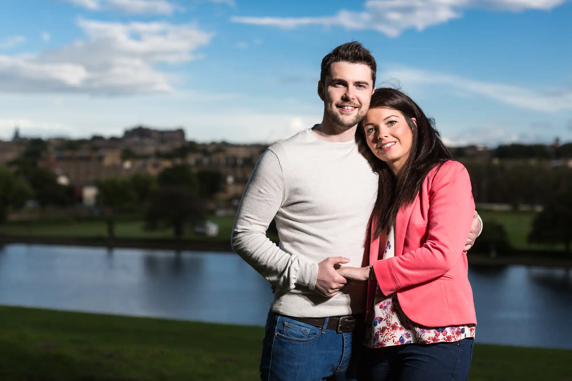 A smiling couple embracing outdoors with a cityscape and river in the background on a sunny day.