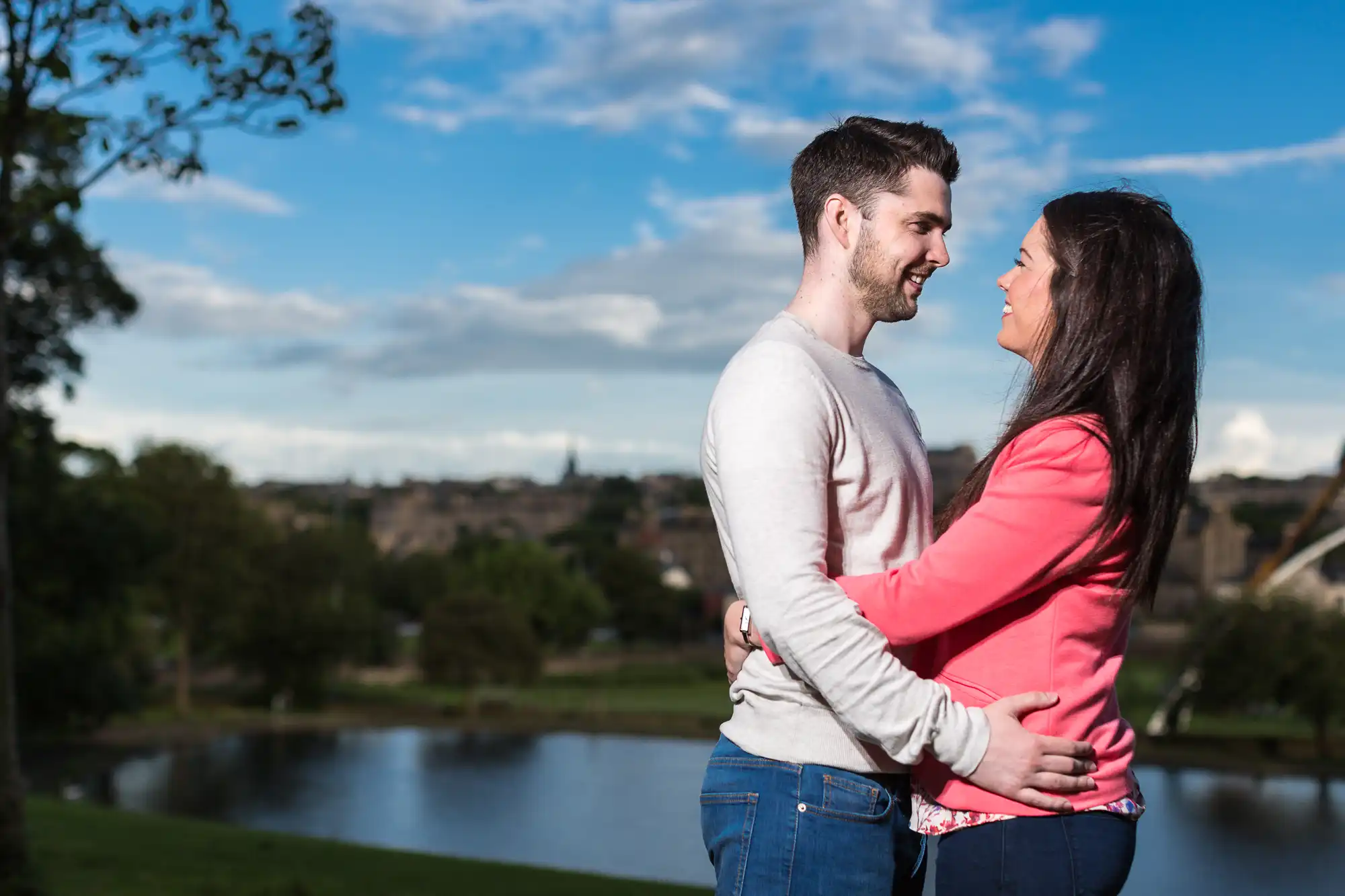 A couple smiling at each other, embracing in a park with a river and cityscape in the background on a sunny day.